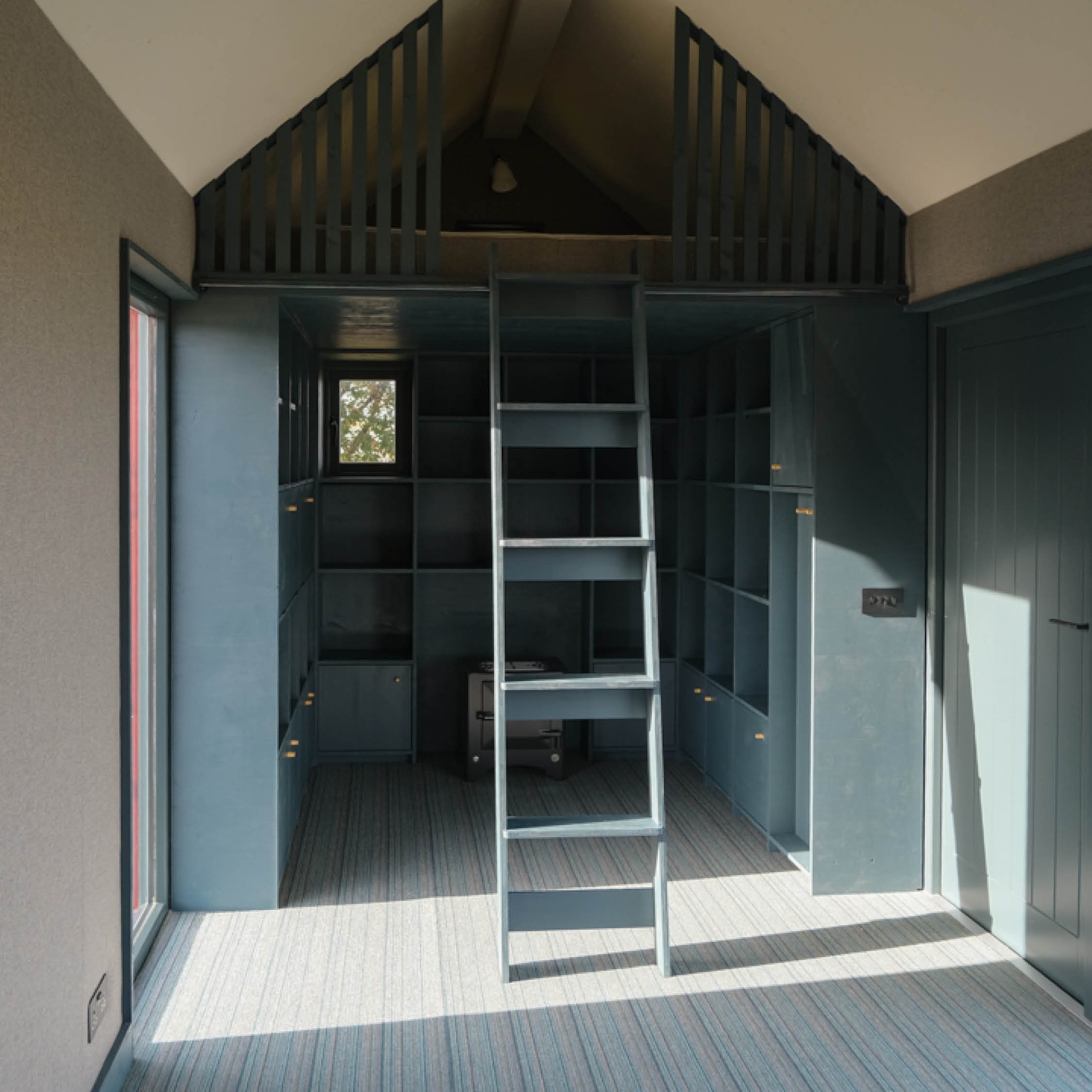 A room with built-in blue cabinets and open shelving, a metal ladder, and a loft area above, with a window and carpeted floor.