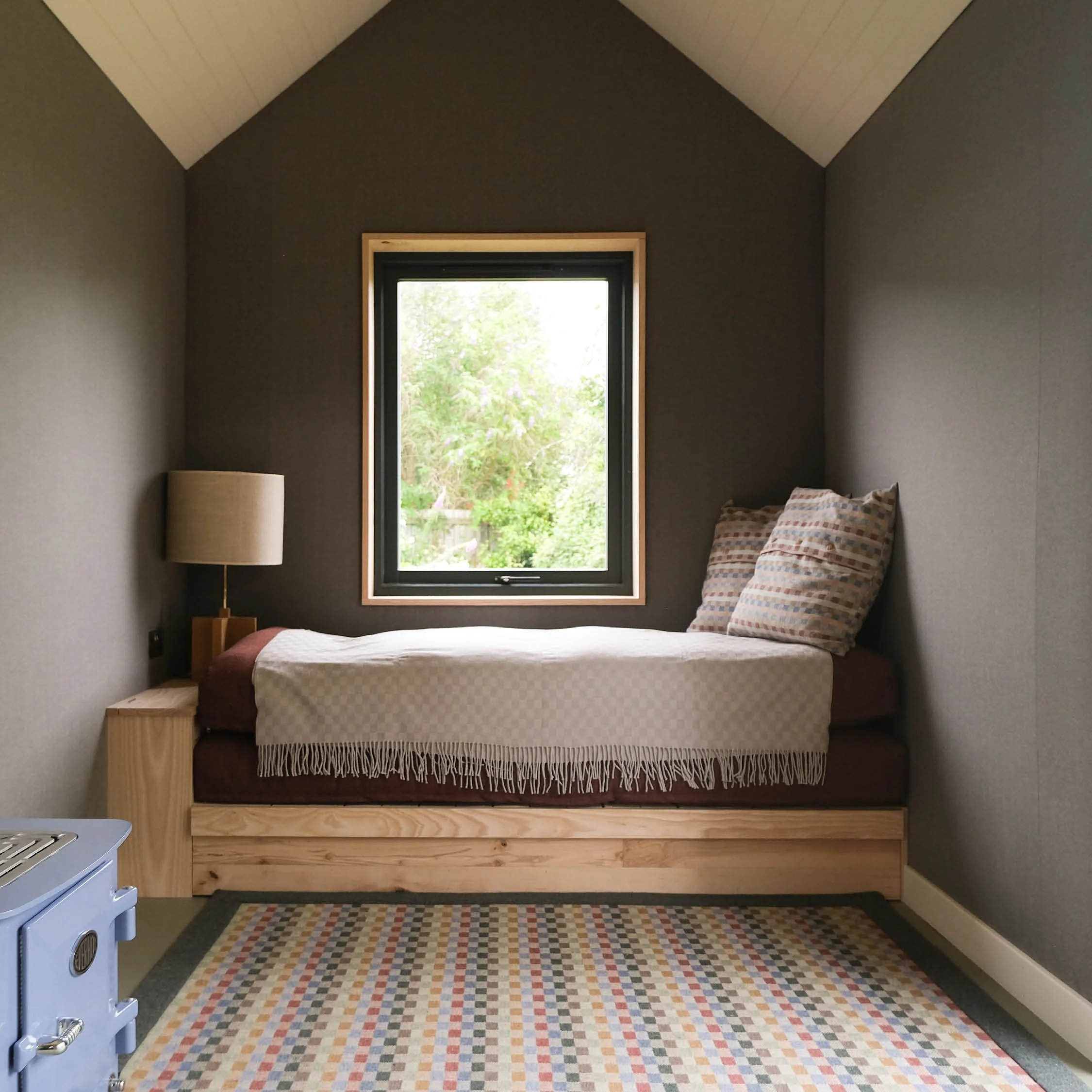 Small bedroom with single bed, patterned pillows, wooden side table, and a window showing greenery outside, beige and gray color scheme.