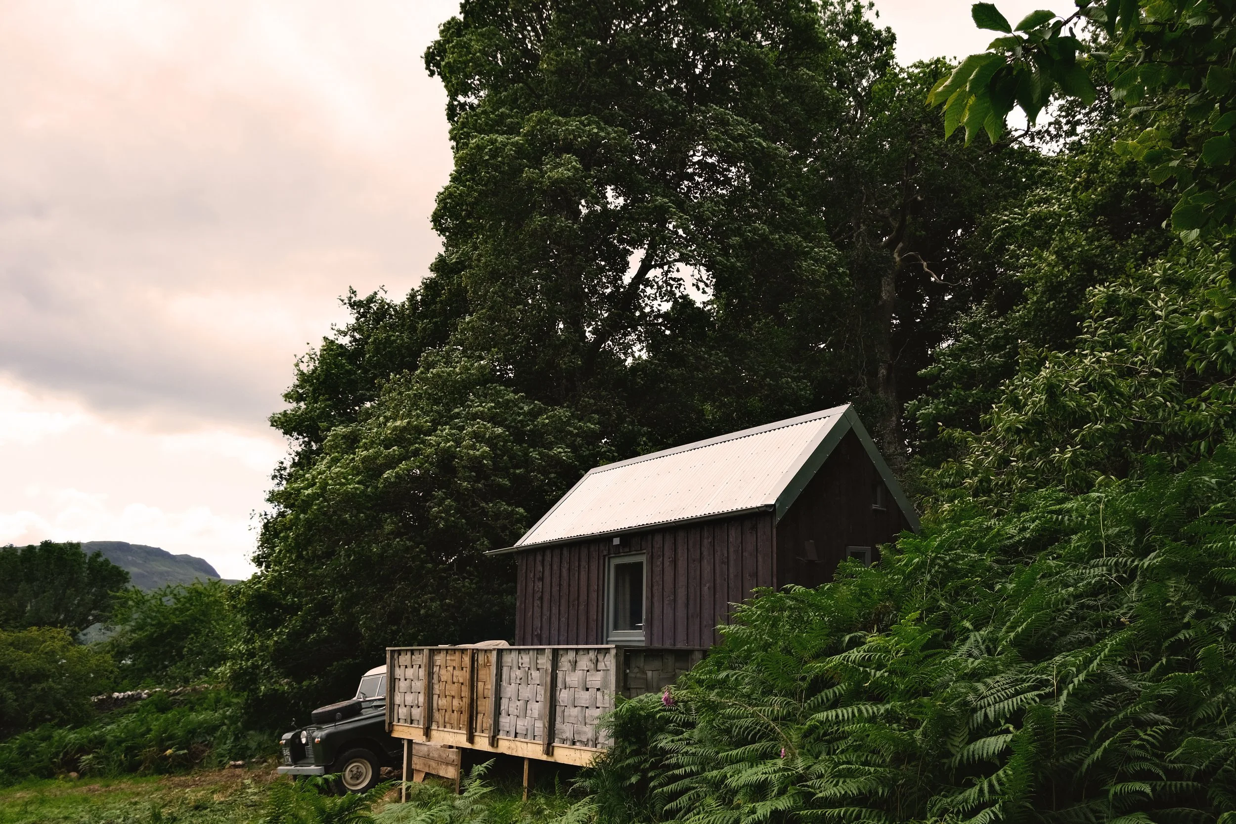 An off-grid hut, surrounded by lush green trees and bushes, and a vintage Landrover parked on the grass nearby.