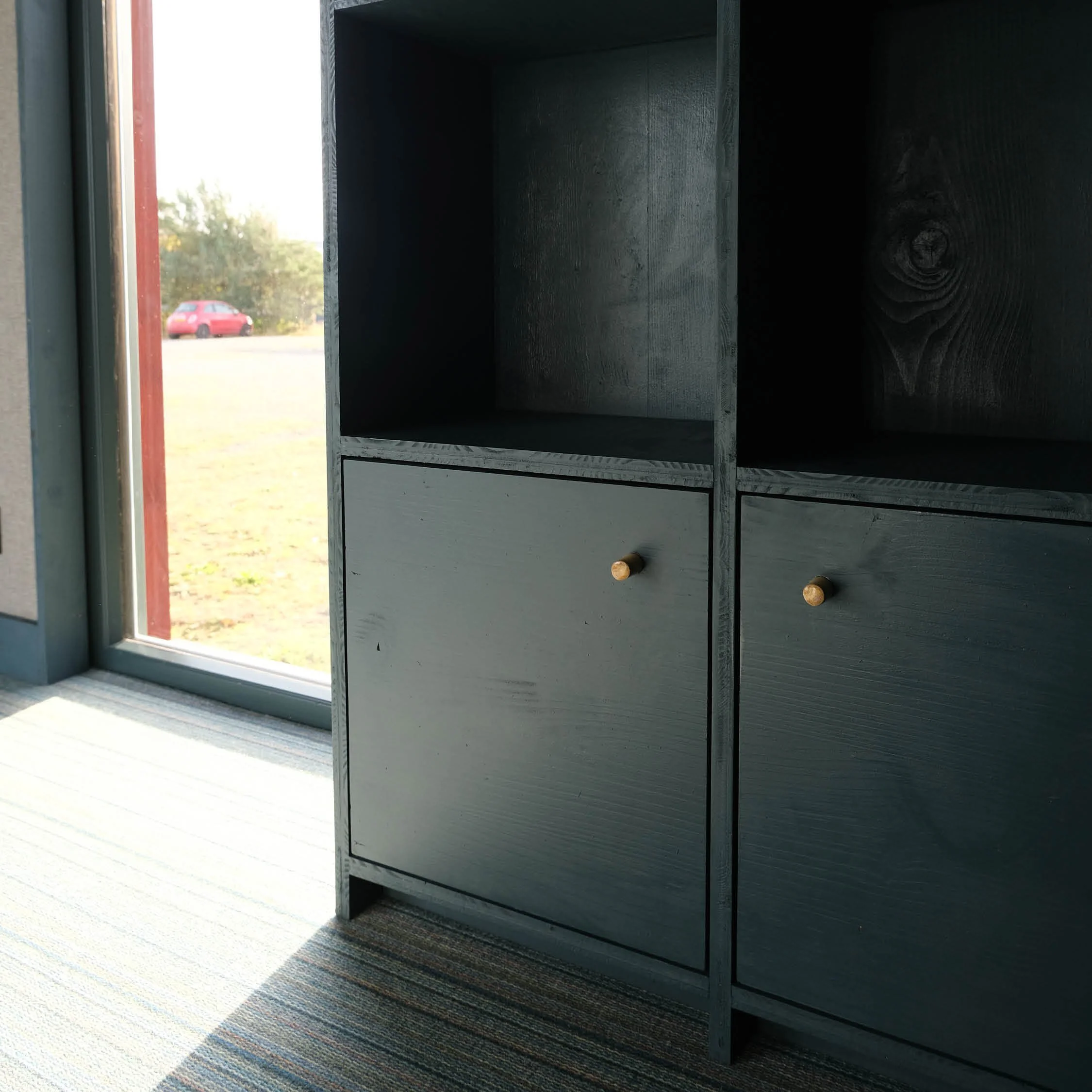 Black wooden shelf unit with four compartments and two small drawers with round ceramic knobs, next to a glass door with an outdoor view.