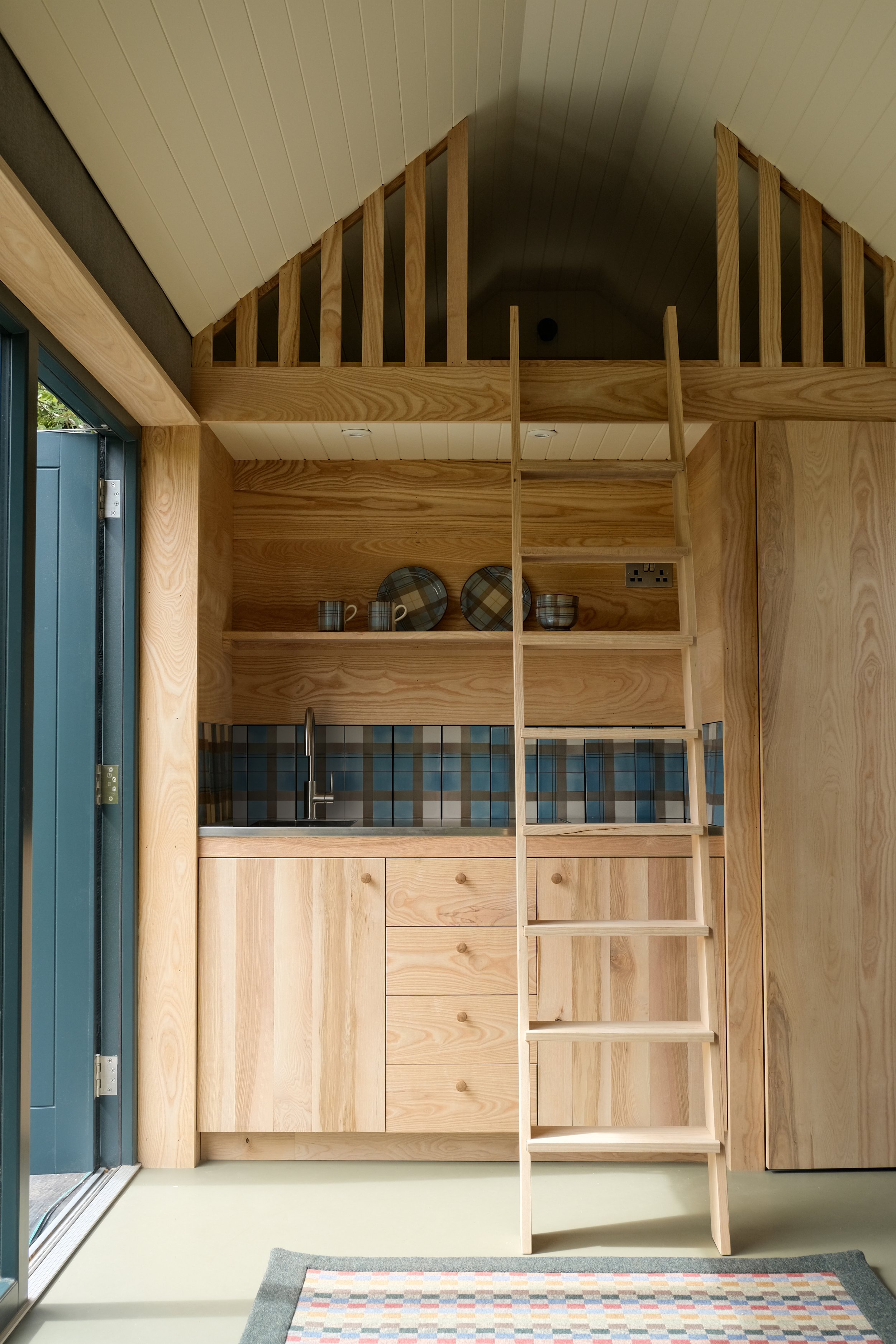 Interior view of a small wooden kitchenette with a ladder leading to an above loft space, featuring a backsplash with tartan tiles, open shelves with plates and bowls, and a small countertop and cabinet.