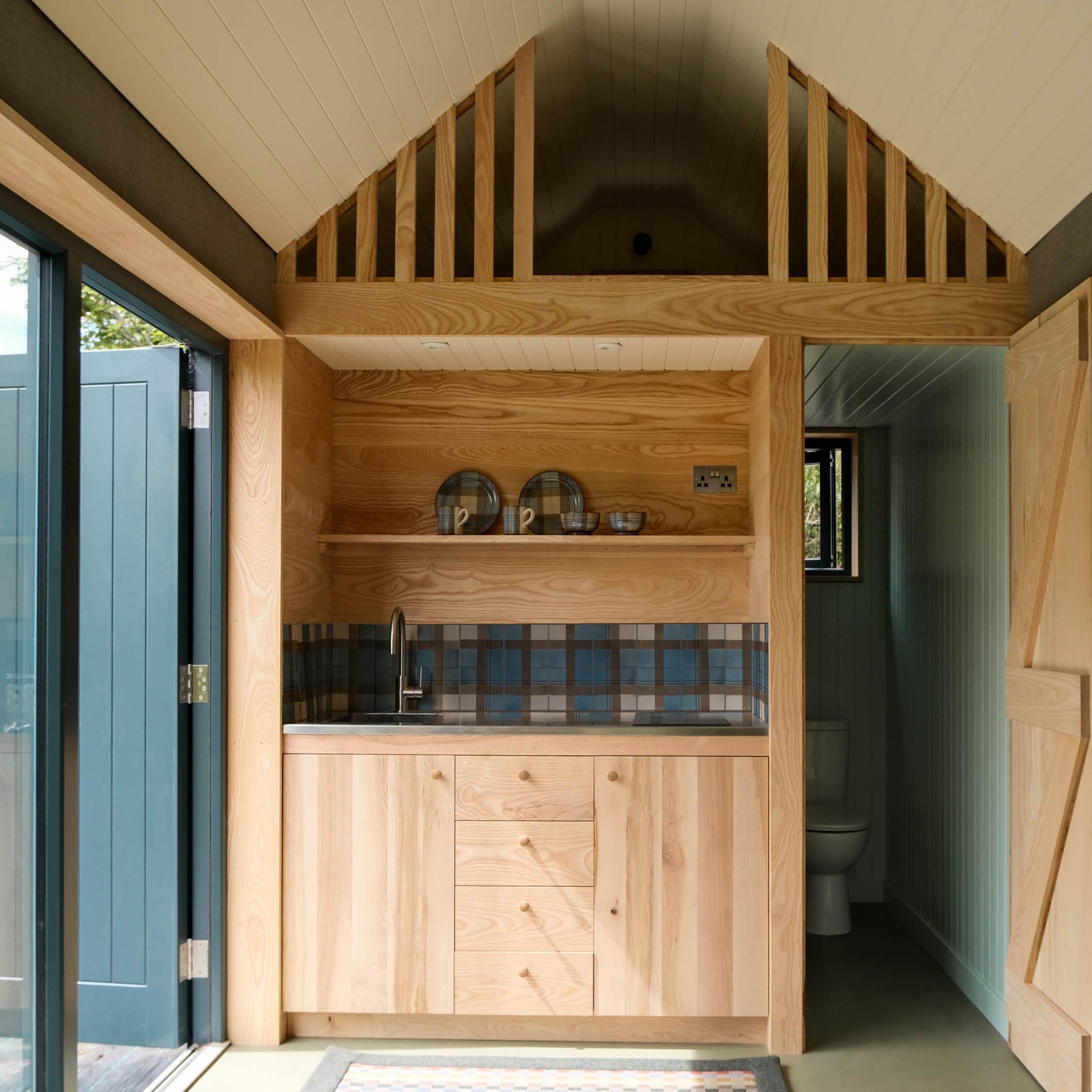 Small kitchen with wooden cabinets, a blue patterned backsplash, and open shelves with dishes. There is a window and a doorway to a bathroom with a toilet visible.