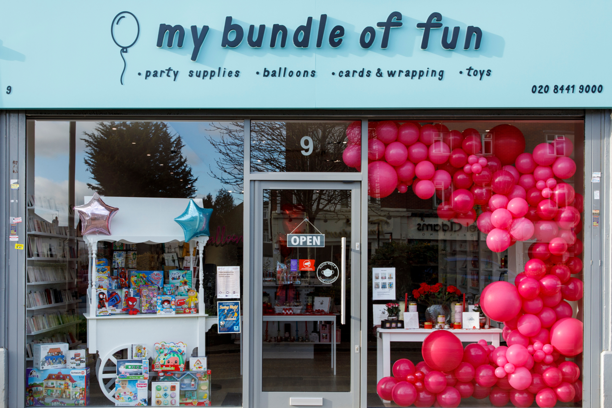 The storefront of 'My Bundle of Fun,' a party supplies, balloons, cards, wrapping, and toys store, decorated with pink and silver balloons, and a display of children's toys in the window.