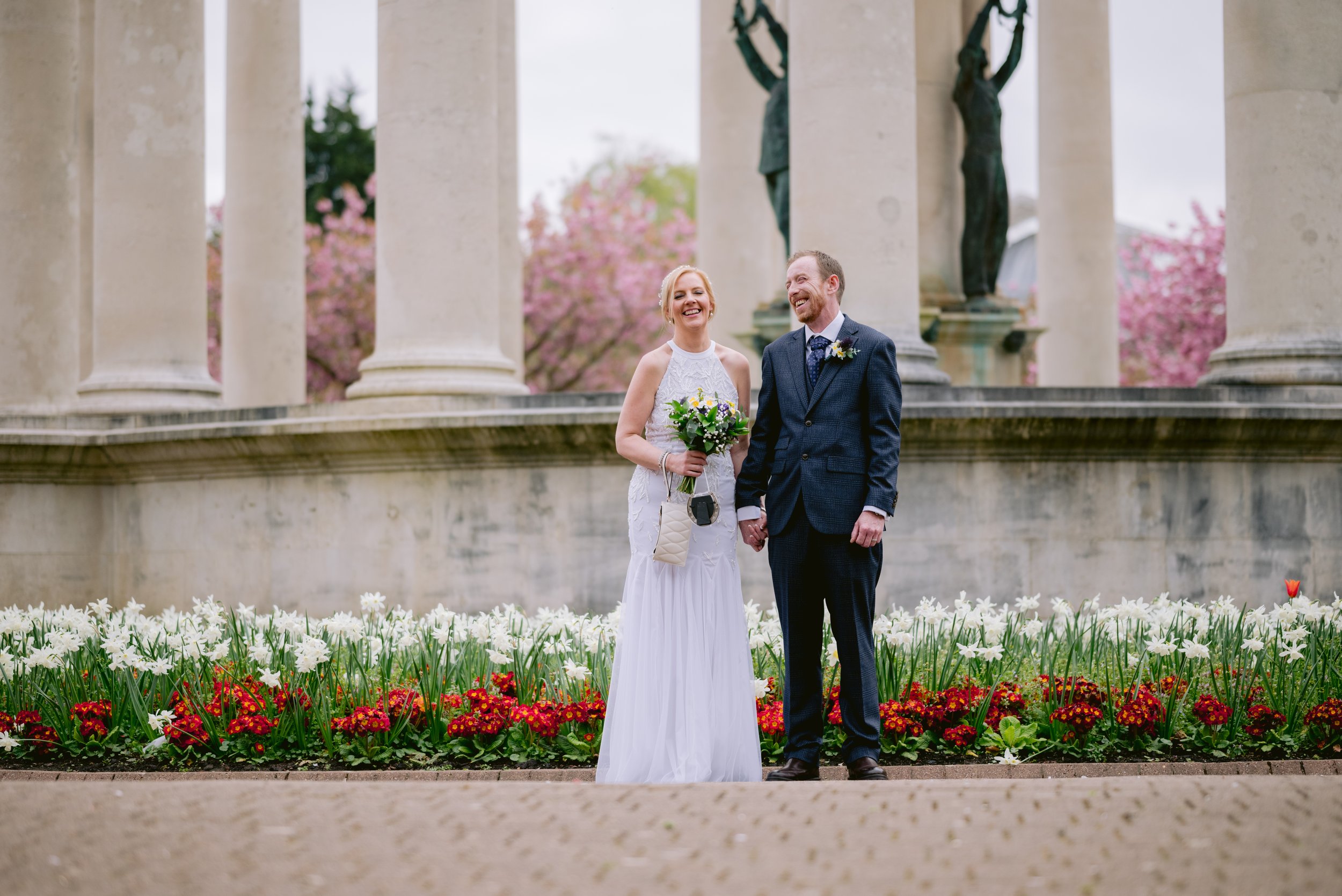 A bride and groom holding hands and smiling, standing in front of a monument with columns and statues, surrounded by white and red flowers and pink blossoms in the background.
