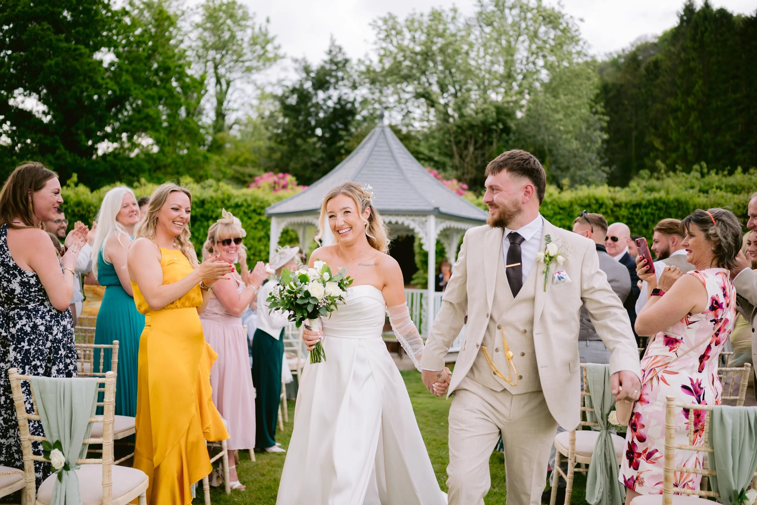 Bride and groom walking hand in hand down the aisle at an outdoor wedding, surrounded by smiling guests clapping and taking photos, with a gazebo in the background.