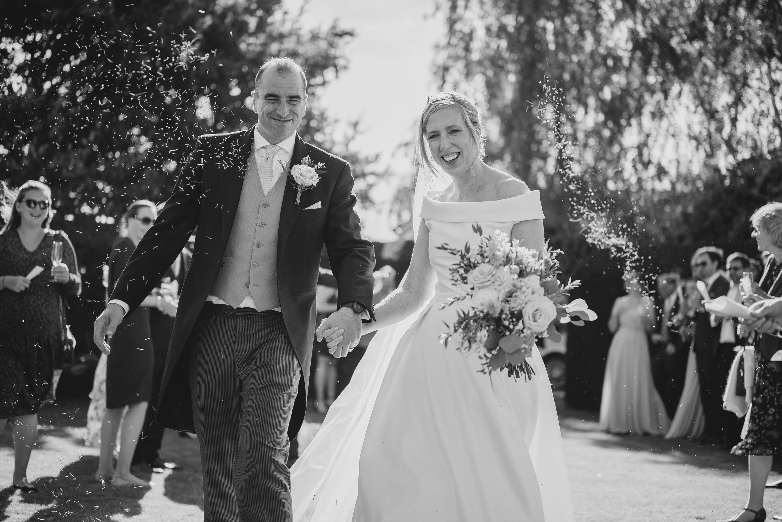 A bride and groom walking hand in hand outdoors after their wedding, surrounded by guests throwing confetti.