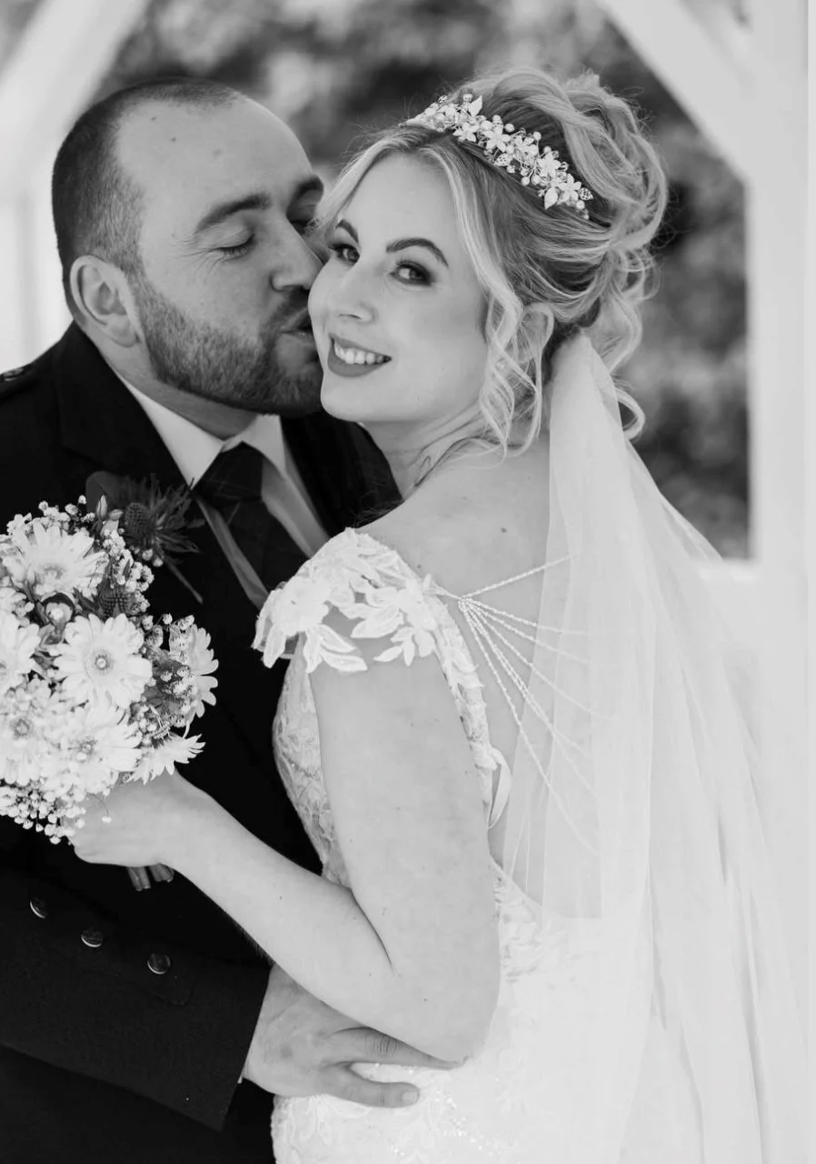 Black and white photo of a bride and groom on their wedding day, close-up, with the groom kissing the bride on the cheek, the bride smiling, wearing a wedding gown and veil, and holding a bouquet of flowers.