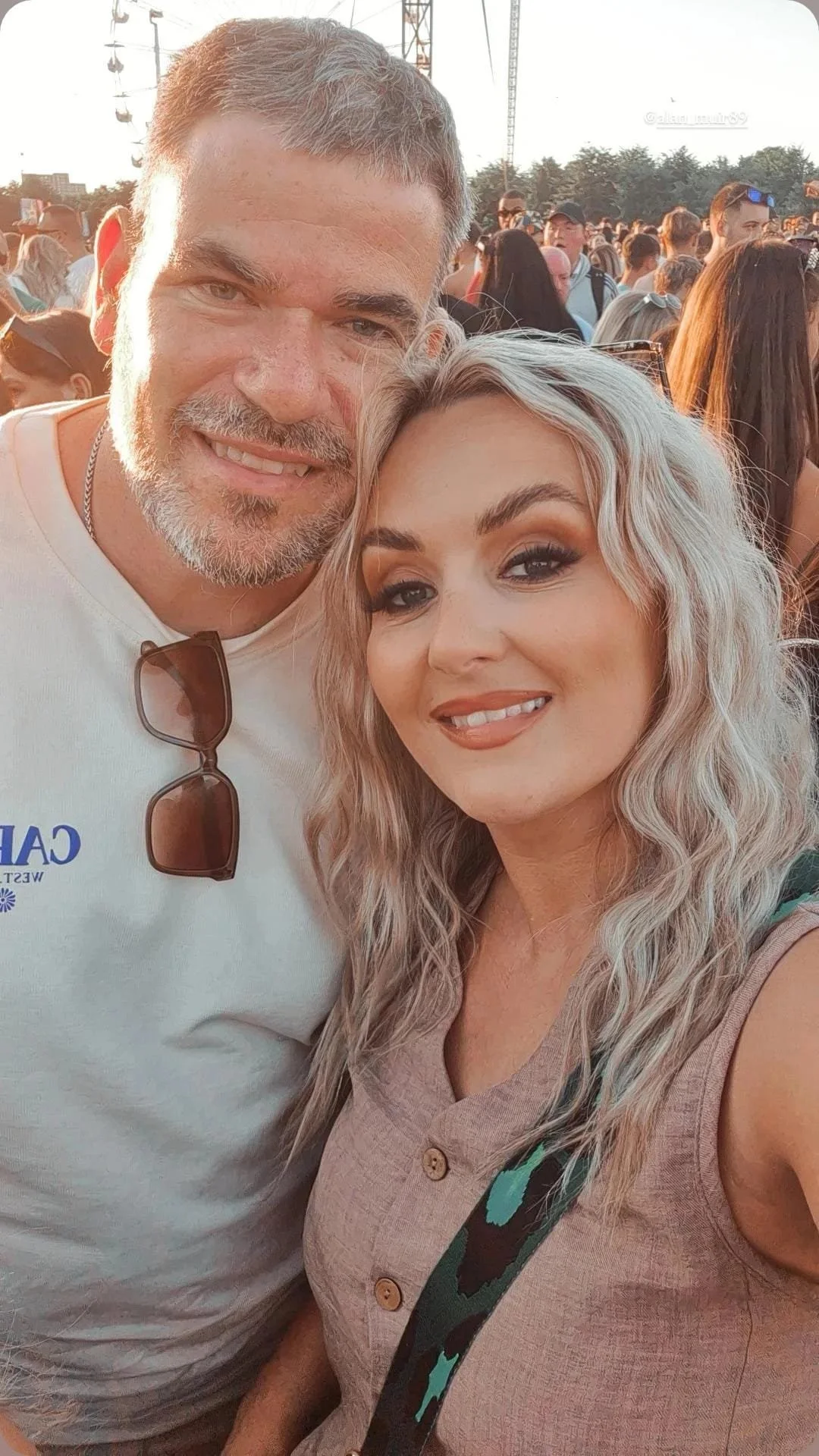 Two people smiling for a selfie at an outdoor event, with many others in the background during sunset.