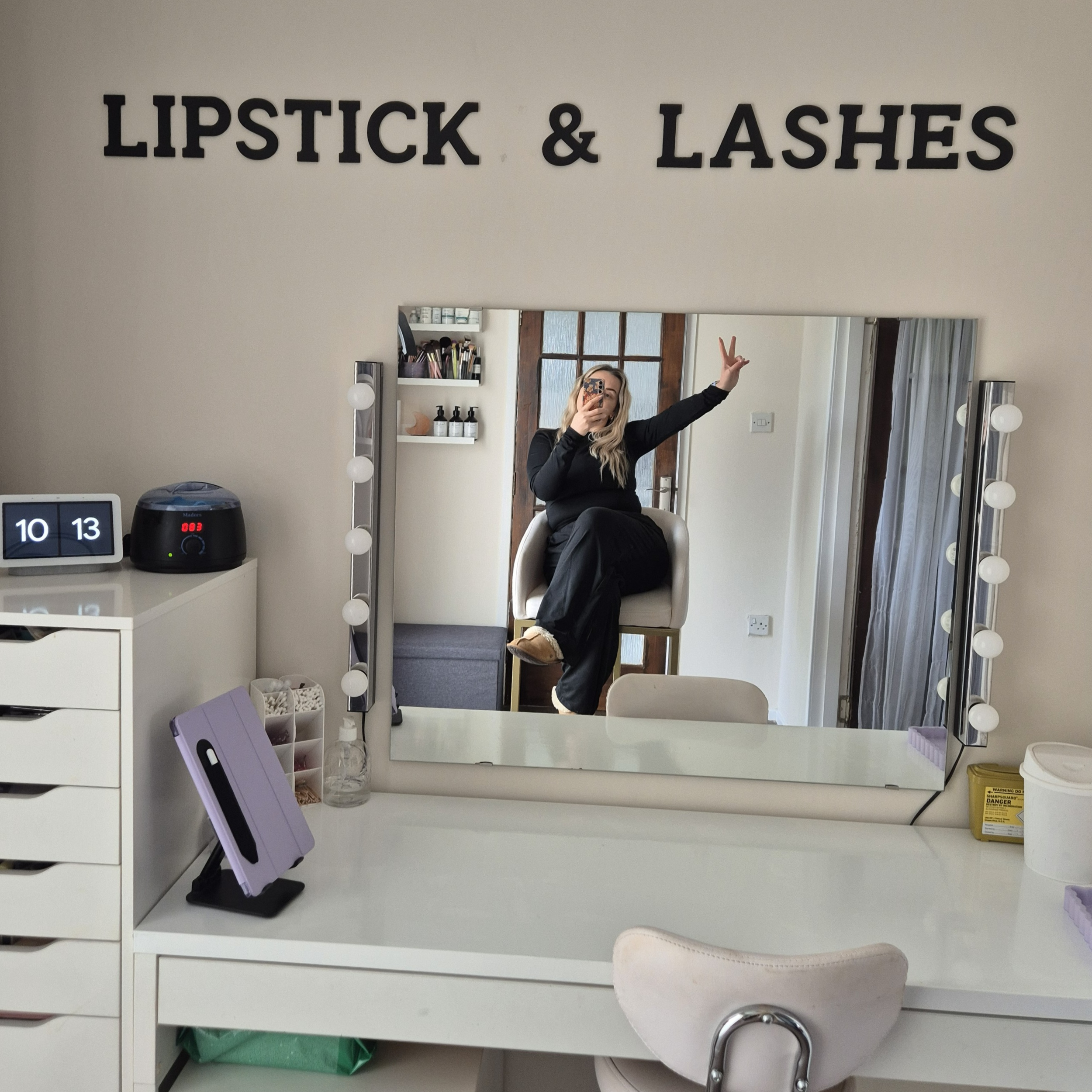 Interior of a beauty salon or makeup studio with a large mirror surrounded by vanity lights. A woman is taking a selfie in the mirror, making a peace sign with her hand, with her legs crossed and sitting on a chair. The top of a white desk and various items including a tablet, clock, and skincare products are also visible.