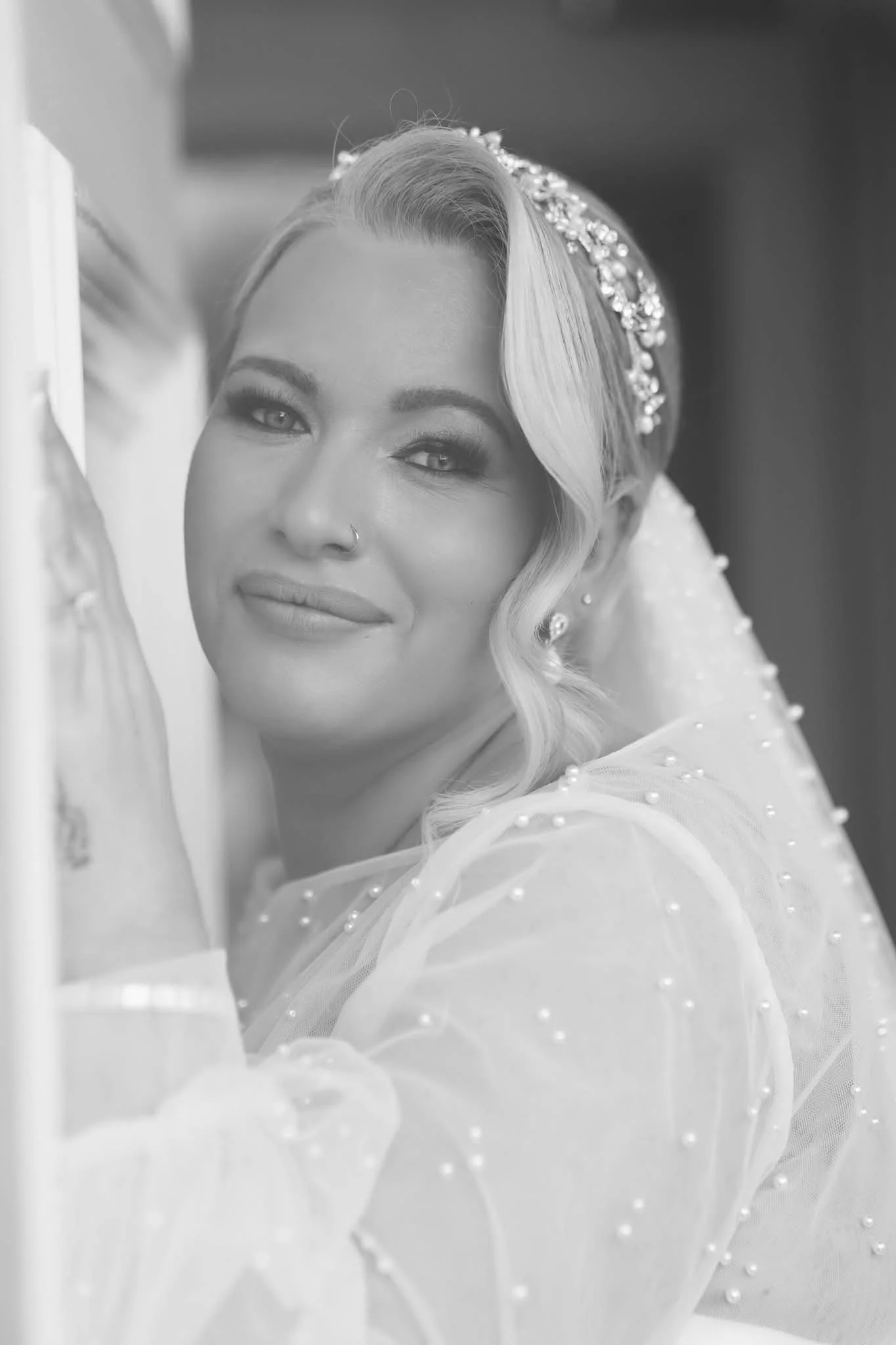 Black and white portrait of a woman with styled wavy hair, makeup, stud earrings, a nose ring, and a beaded veil or headpiece, looking at the camera with a soft smile.