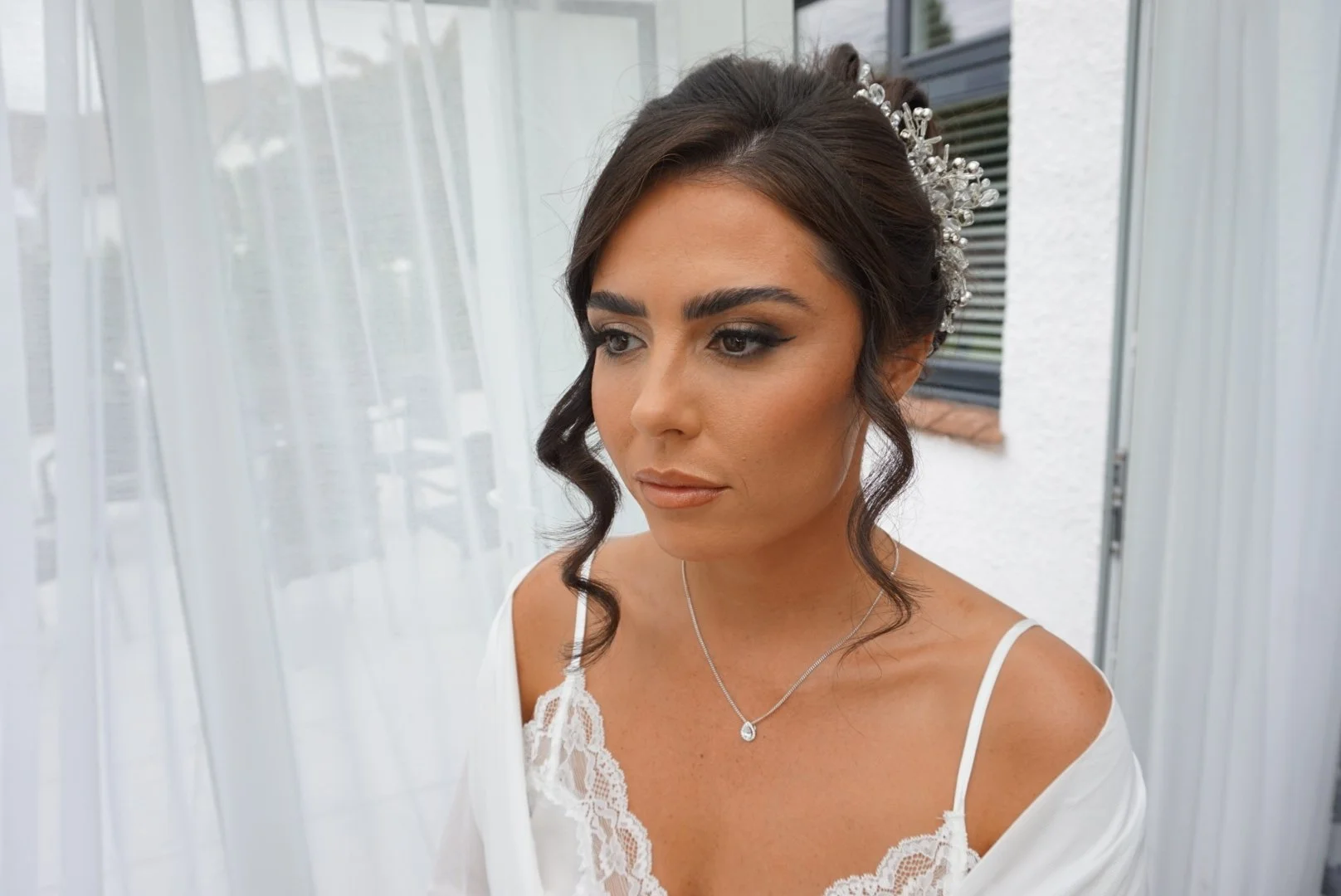 A woman with dark hair styled in loose curls, wearing a decorative hair accessory, a white lace dress, and a silver necklace, standing near a window with white curtains.