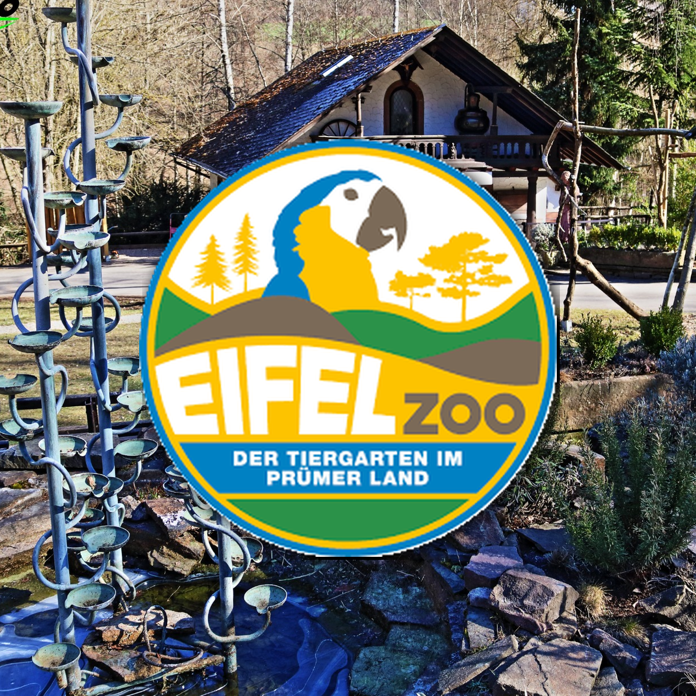 Das Logo des Eifelzoos, ein bunter Kreis mit einem blauen Vogel, Bergen, Bäumen und dem Text 'Eifel Zoo, Der Tiergarten im Prümer Land', vor einem Tiergehege mit Spielgeräten in einem Waldpark.Eifel Zoo Wildpark Tiere Tierpark Lünebach Park Plan Info