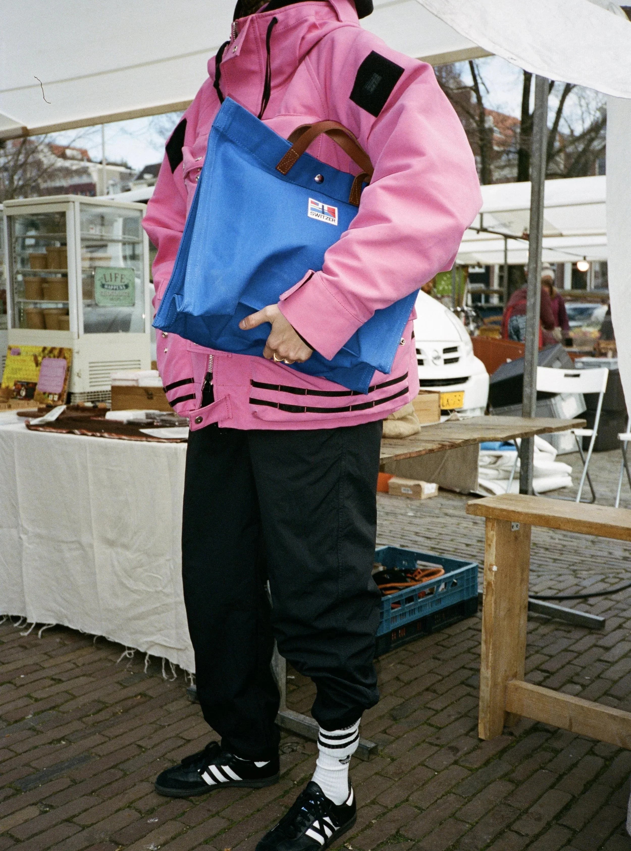 Person wearing a pink jacket, black pants, white socks with black stripes, and black sneakers, holding a blue bag at an outdoor market with tables and other people in the background.