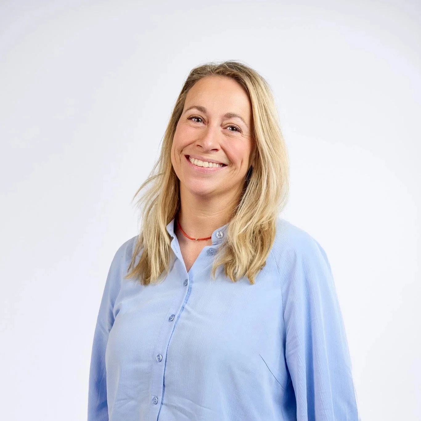 Vera van Ham - Zilver Vitaal - A woman with long blonde hair smiling, wearing a light blue button-up shirt, indoors with blurred background of large windows and modern decor