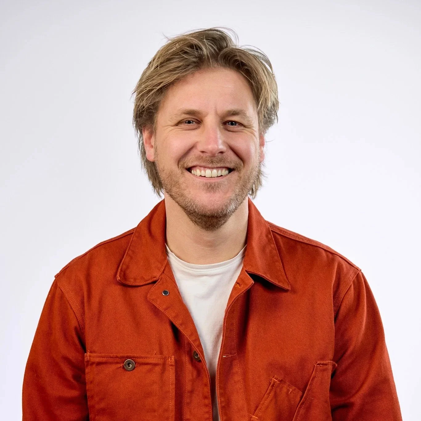 Close-up of a smiling man with blond hair, blue eyes, and a beard, wearing a green shirt, against a white background.