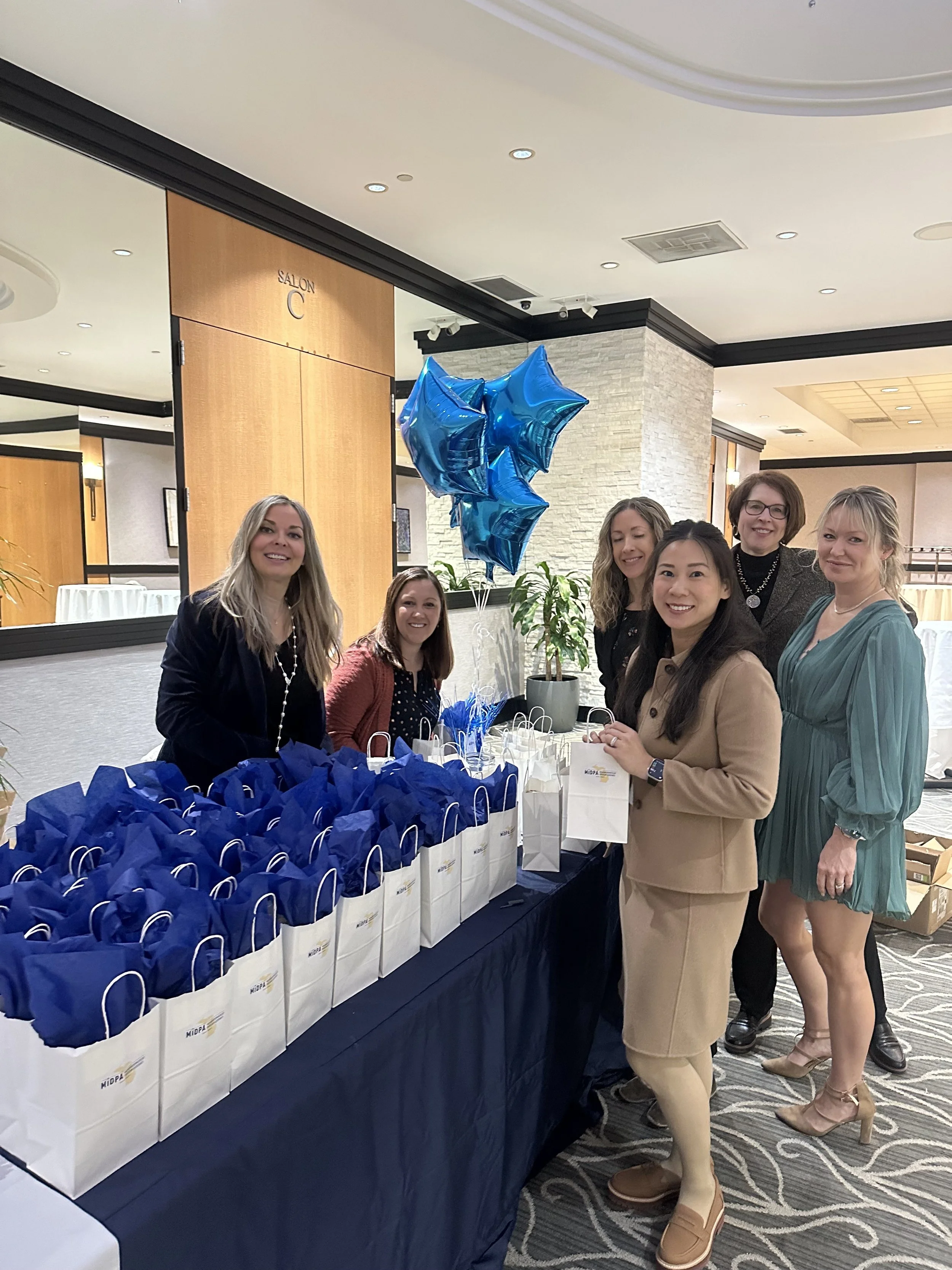 A group of six women standing behind a table with gift bags at a formal event, some holding white and blue balloons.