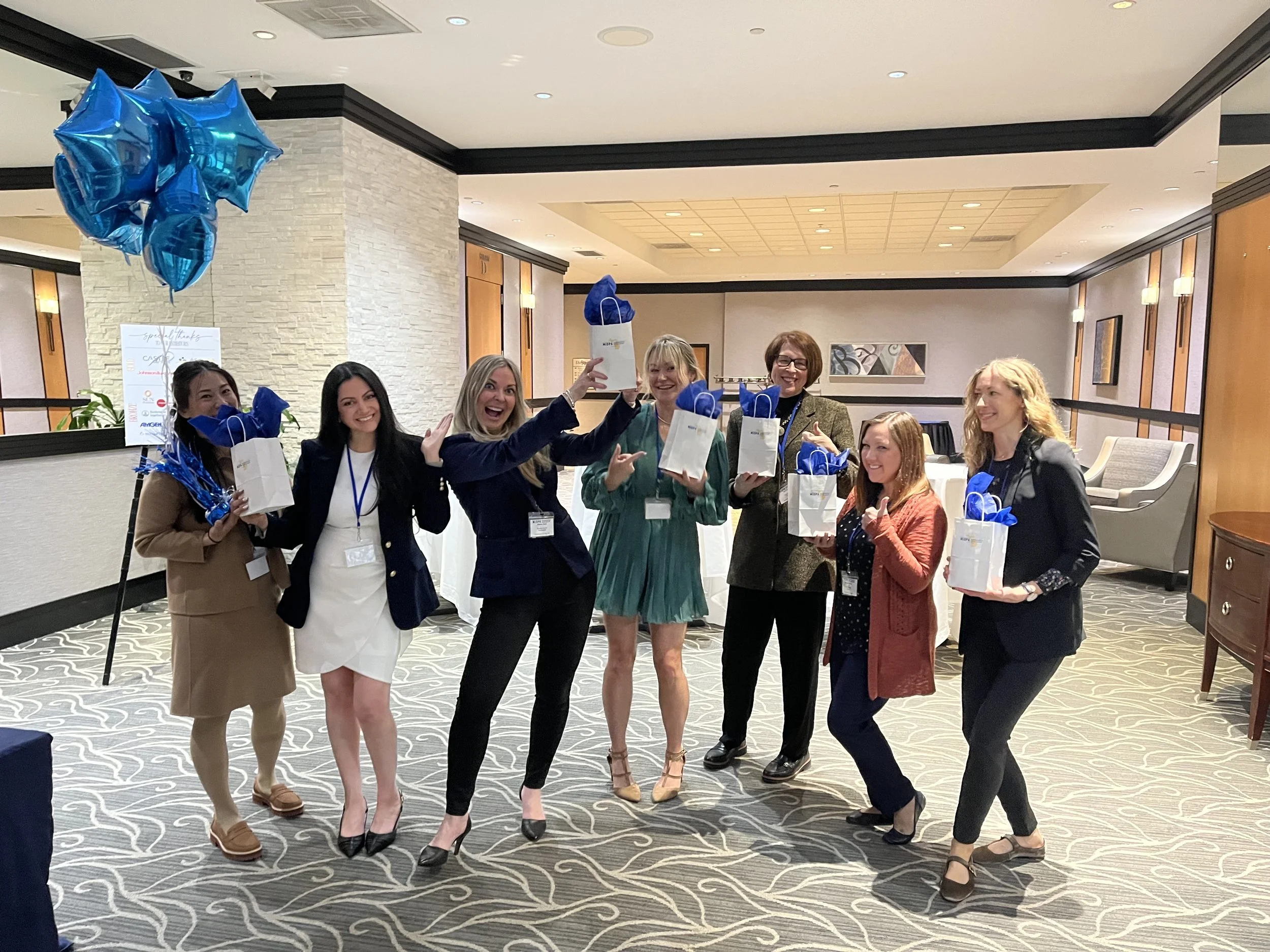 Group of six women celebrating indoors at a conference or event, holding white gift bags with blue tissue paper and smiling.