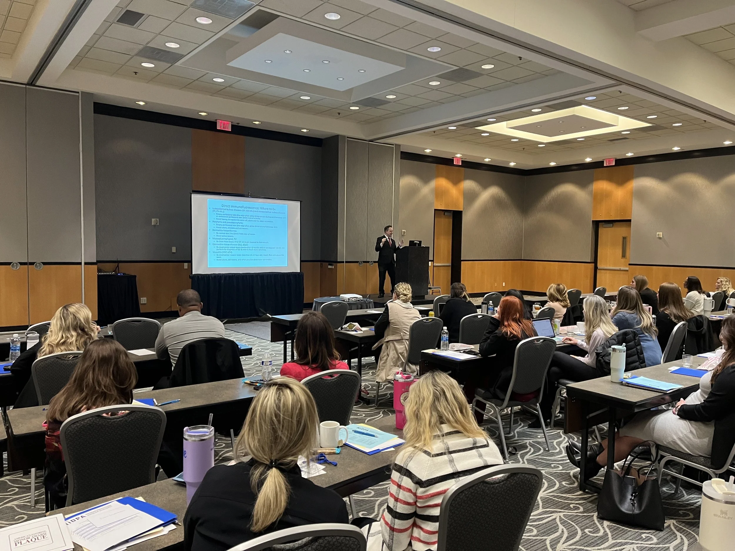 A speaker in a suit giving a presentation at a conference room with seated attendees, bottle water, notebooks, and a large screen displaying a slide.