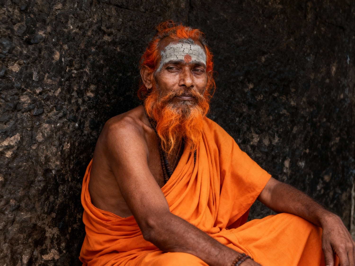 Sâdhu en robe orange, cheveux et barbe teints au curcuma, adossé à la roche — Inde