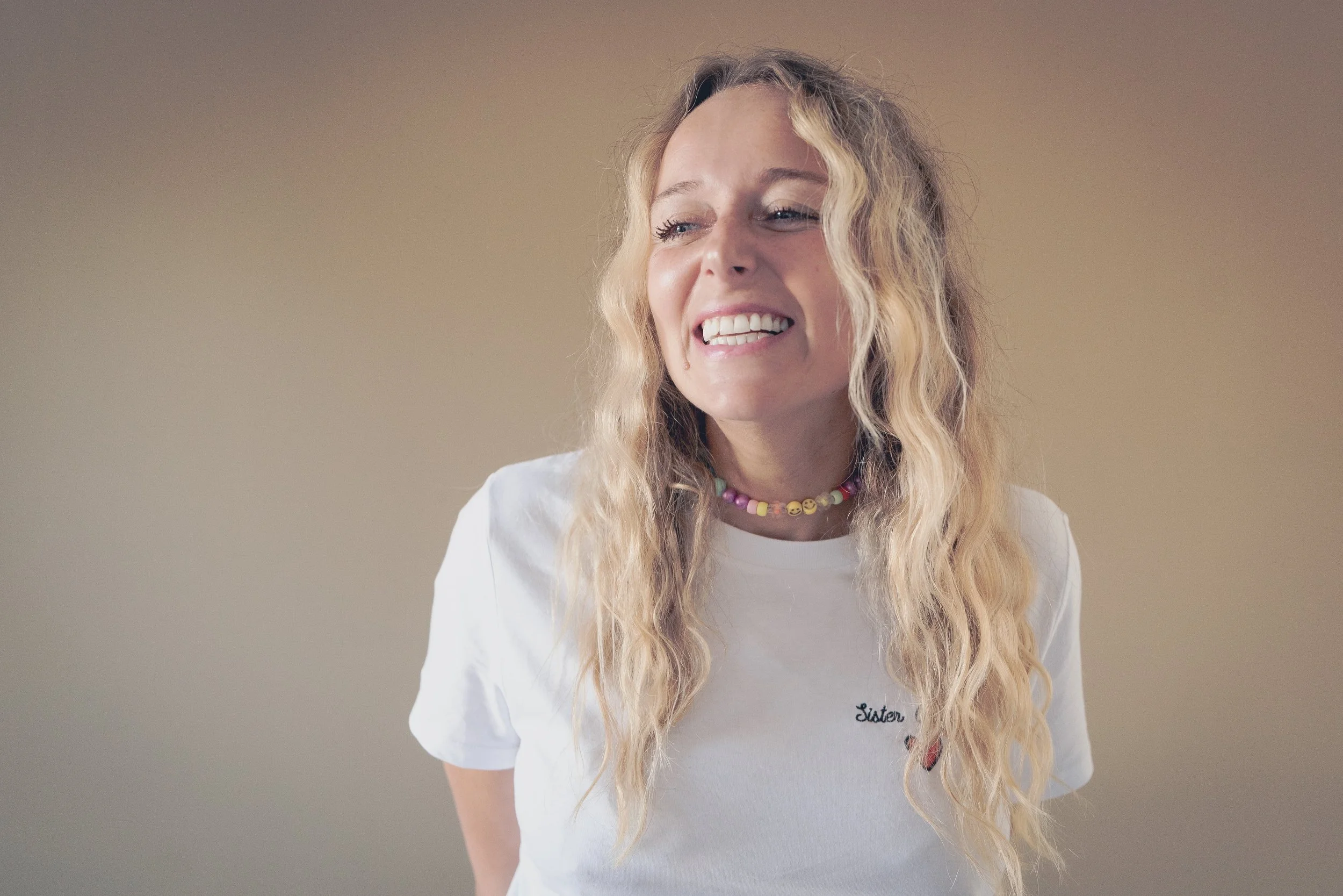 Jeune femme avec une expression de douleur ou de frustration, portant un t-shirt blanc avec le mot 'Sister' et un collier coloré, sur un fond neutre.