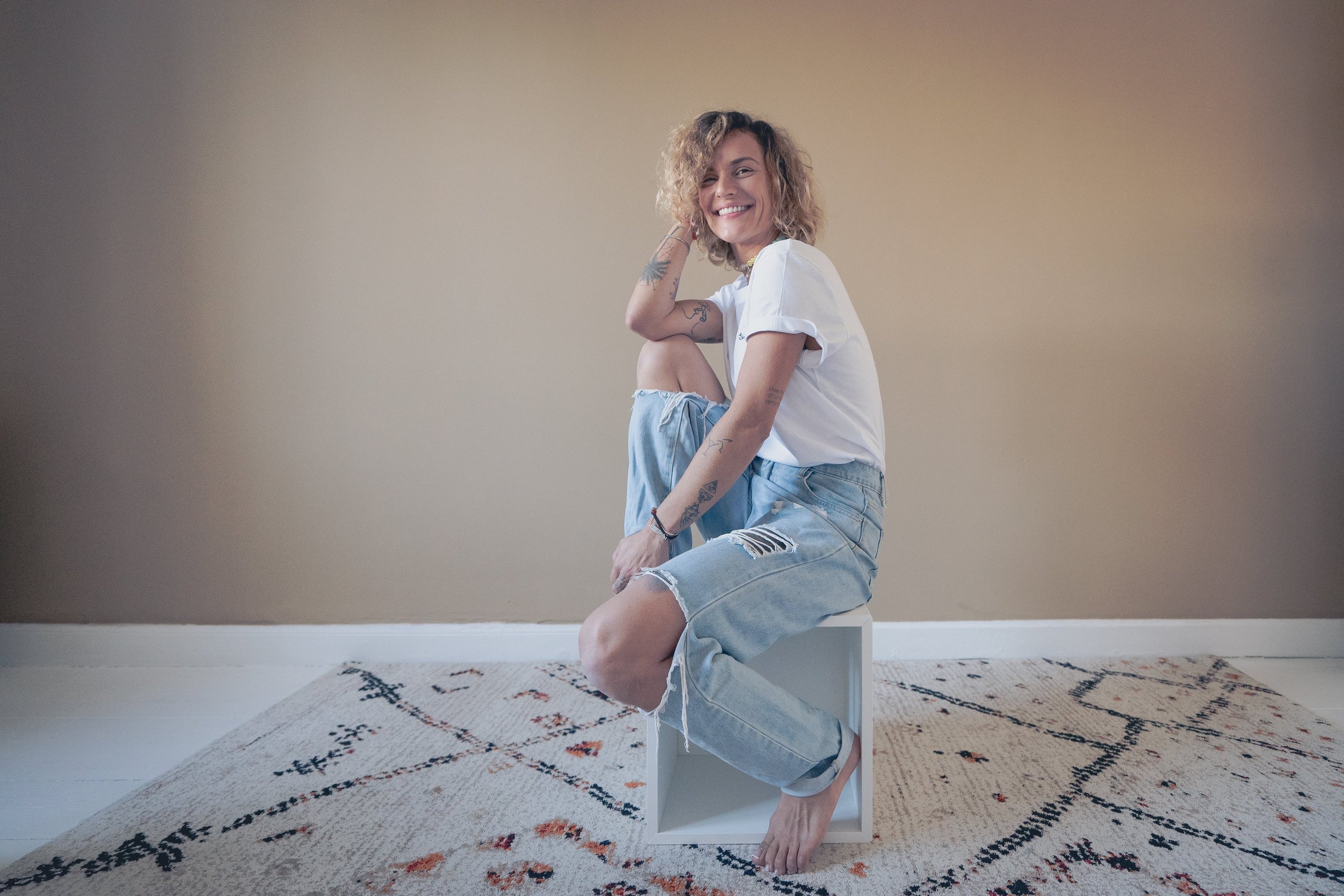 Femme souriante aux cheveux bouclés, portant un t-shirt blanc et un jean déchiré, assise sur une boîte en bois sur un tapis beige avec un motif géométrique, dans une pièce avec un mur beige.