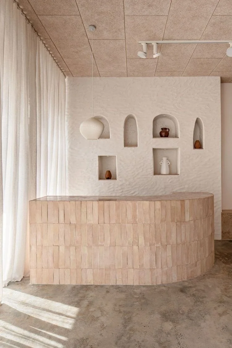 Minimalist curved reception desk with a textured white wall behind it, built-in decorative shelves, and beige curtains on the left side. The ceiling has a brown tile finish and a track light fixture
