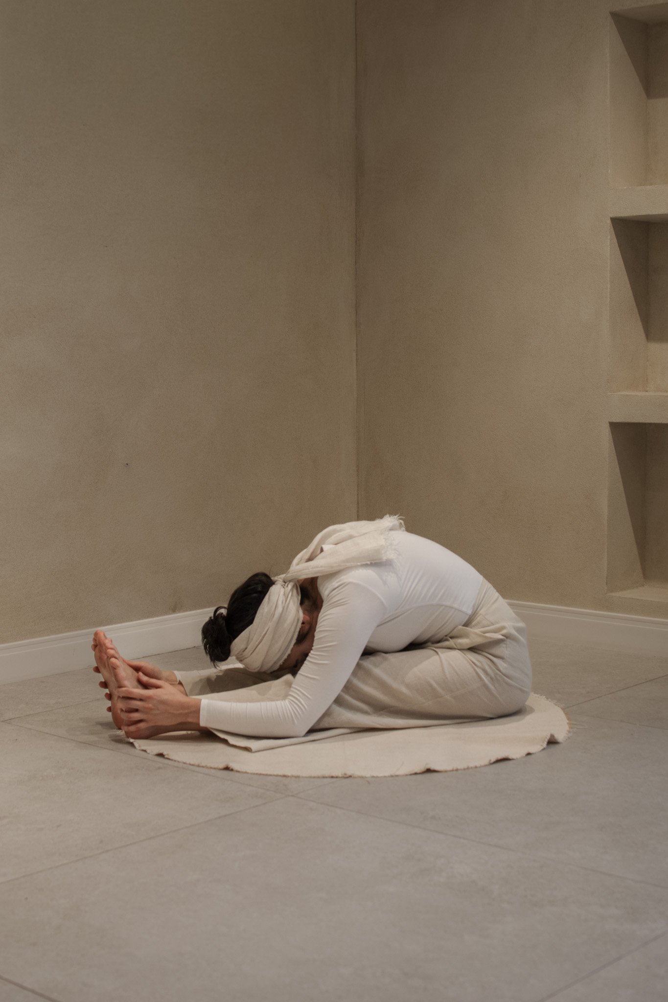 Person dressed in white in a seated forward bend yoga pose on a white rug in a neutral-colored room.