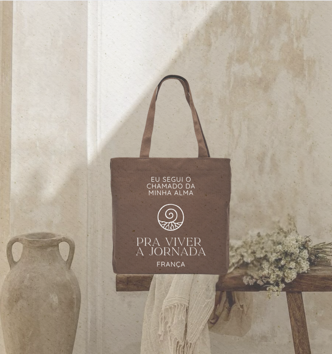 Brown tote bag with Portuguese text and spiral logo hangs on a wall in a minimalistic room with a large ceramic vase, a wooden table, a cloth, and a bunch of white flowers in the background.