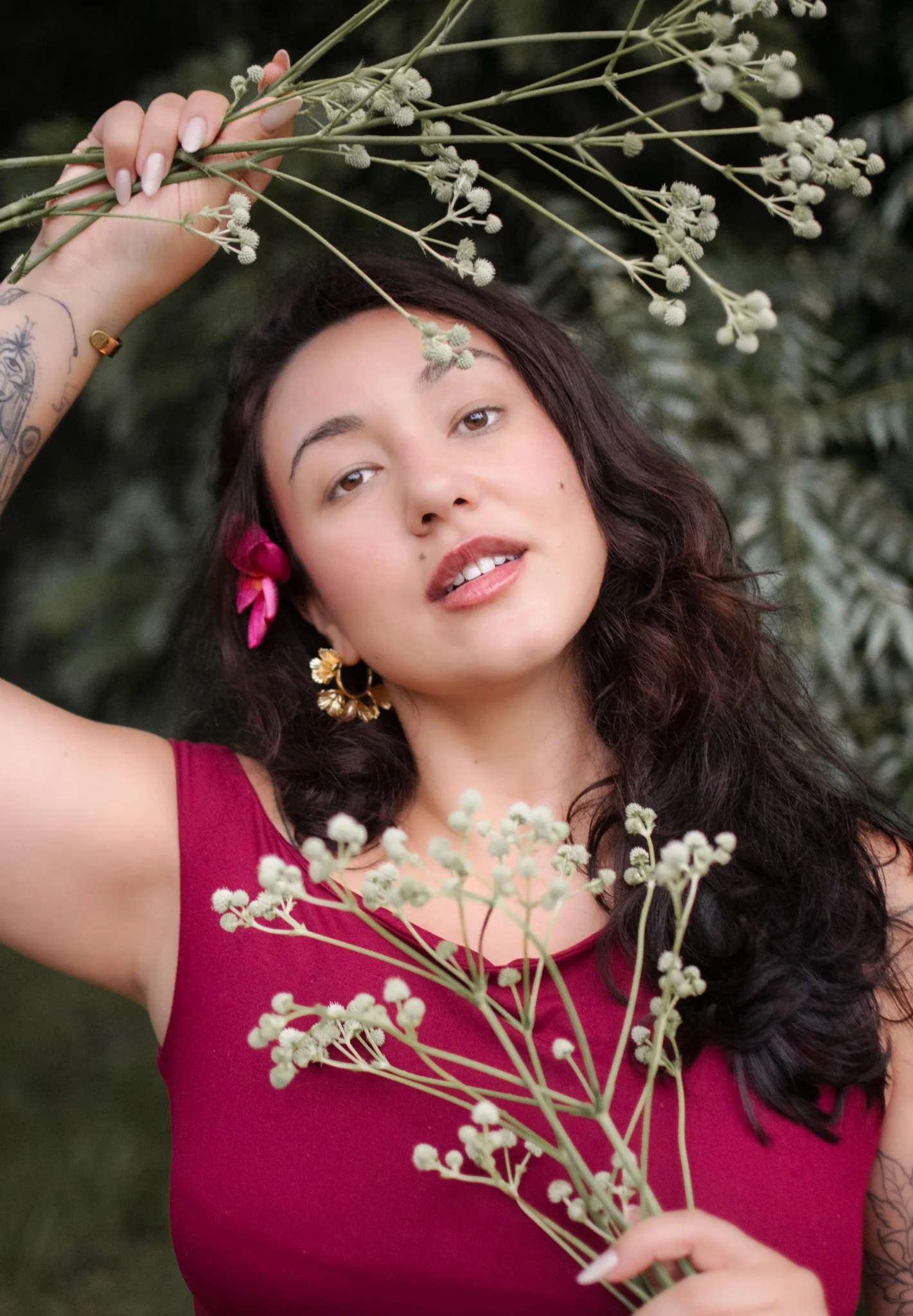 A woman with long dark hair, wearing a red sleeveless top, holding white flowers, standing outdoors with greenery in the background.