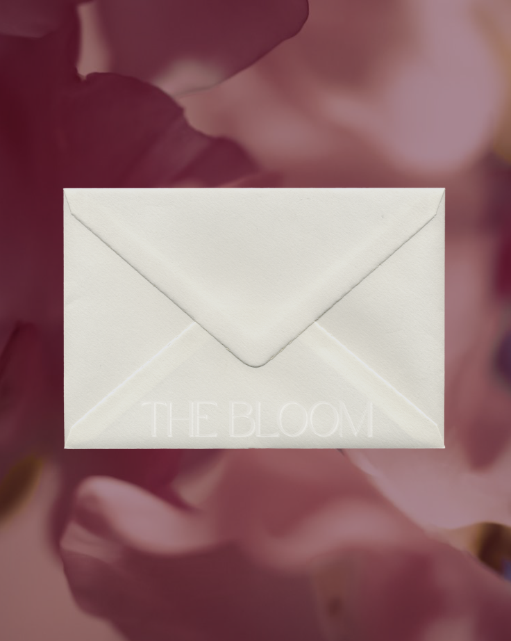 A white envelope with the words 'THE BLOOM' embossed on it, placed against a blurred pink and purple floral background.