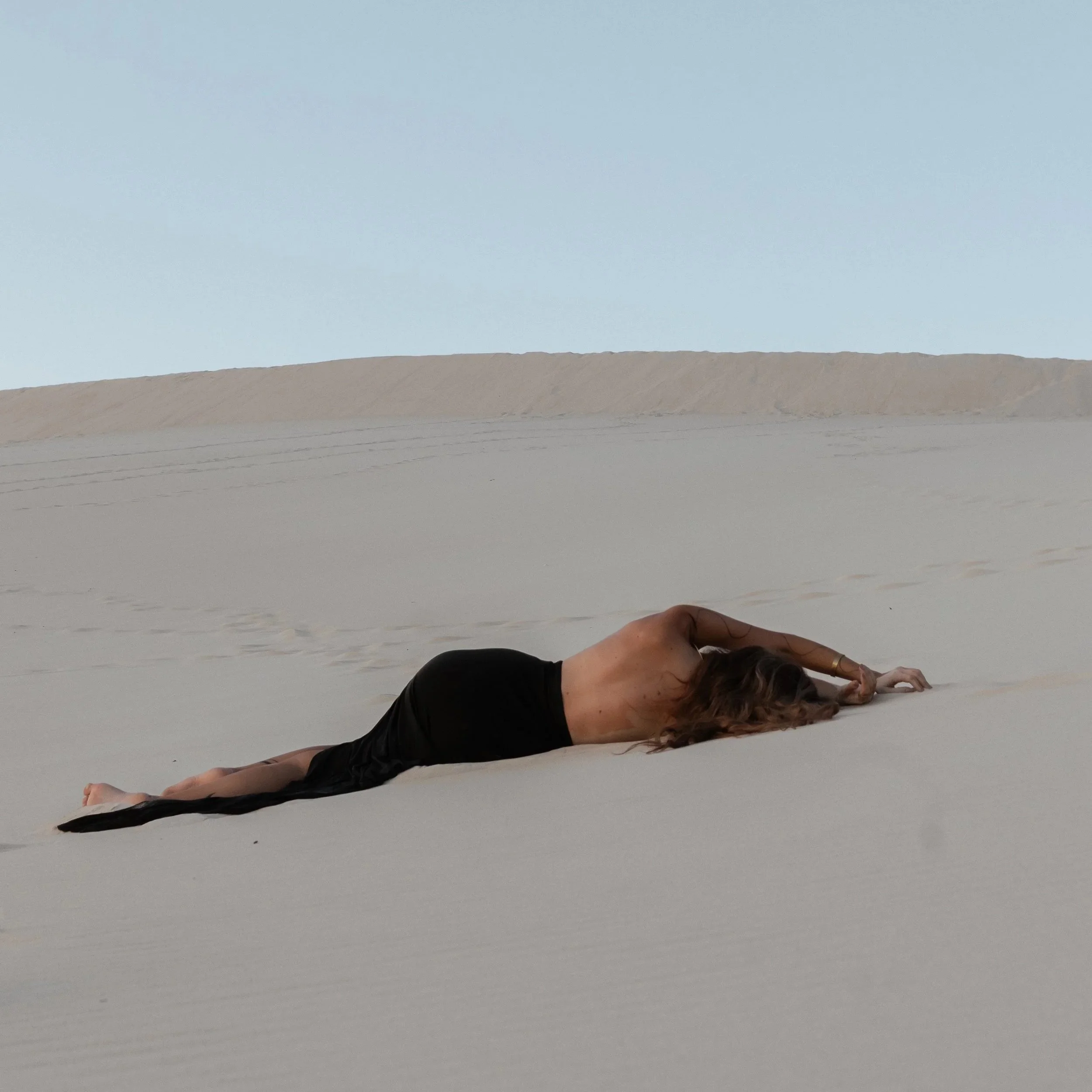 A person lying flat on their stomach on a sandy desert with arms stretched out forward and head turned to side, wearing black pants and topless, with a vast expanse of sand dunes and pale sky in the background.