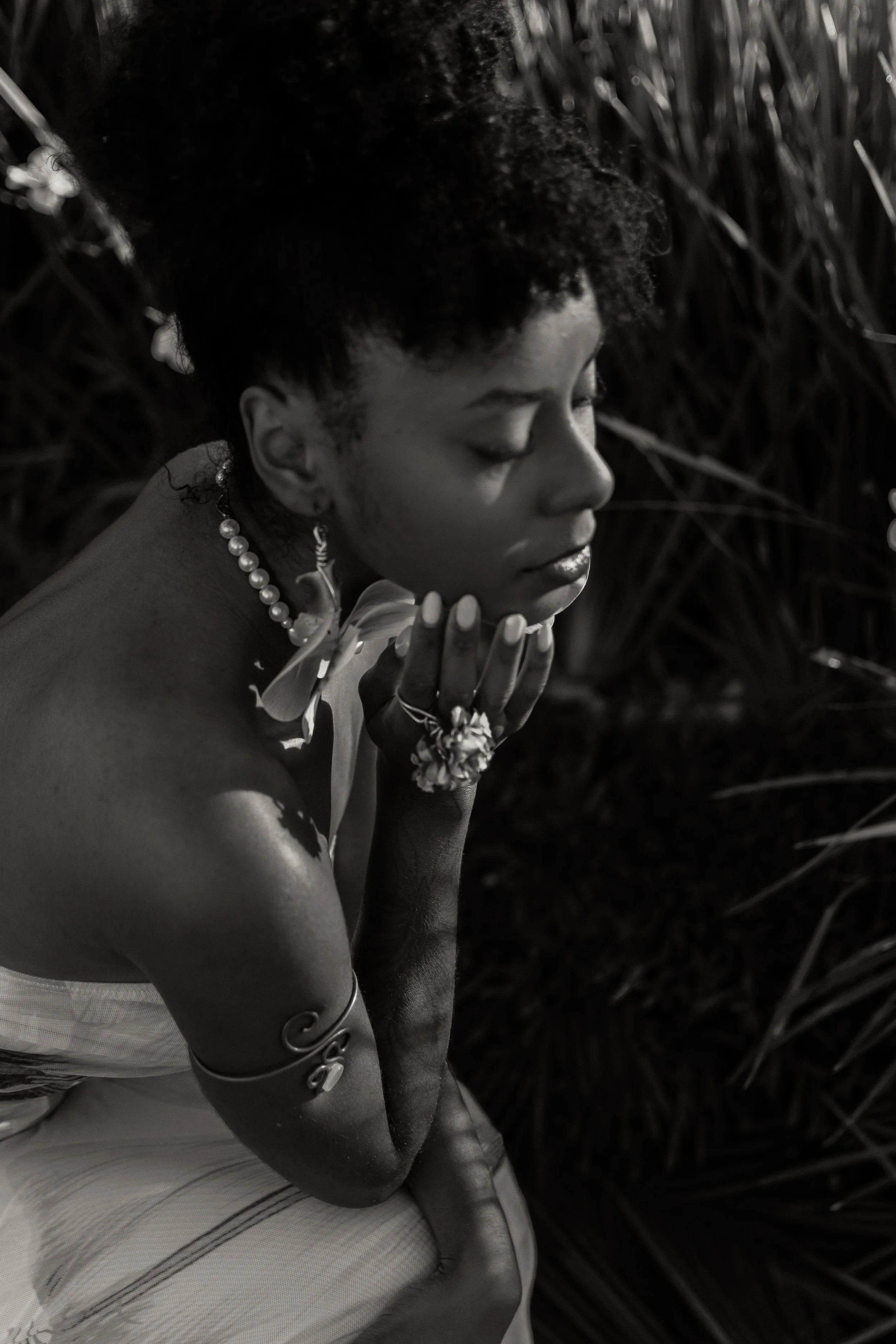 Black and white photo of a woman with curly hair, jewelry, and tribal tattoos, sitting with her eyes closed and hand on her chin in a natural outdoor setting.