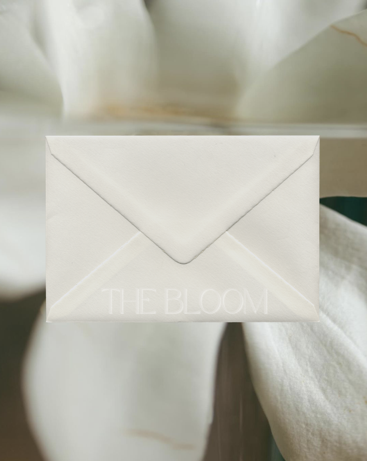 A white envelope with embossed text saying 'THE BLOOM' on a cream-colored surface.
