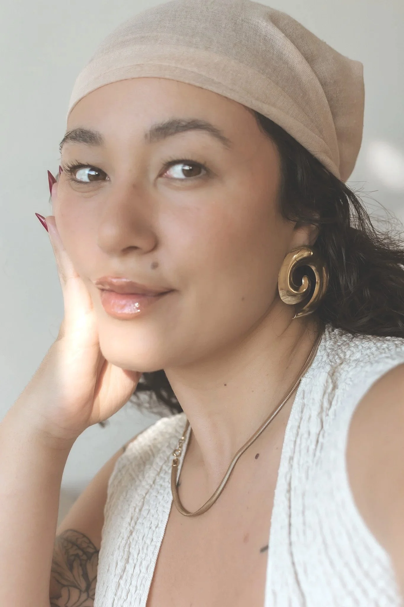 Close-up portrait of a woman with dark curly hair wearing a beige headscarf, gold swirl earrings, and a white textured sleeveless top. She has light makeup, glossy lips, and a small beauty mark above her lip. She is resting her face on her hand and looking into the camera.