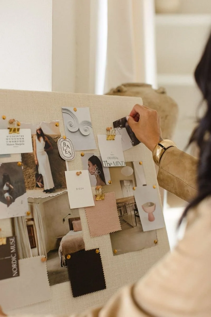 A woman pinning photos and fabric swatches to a fabric-covered board with pushpins in a living space.