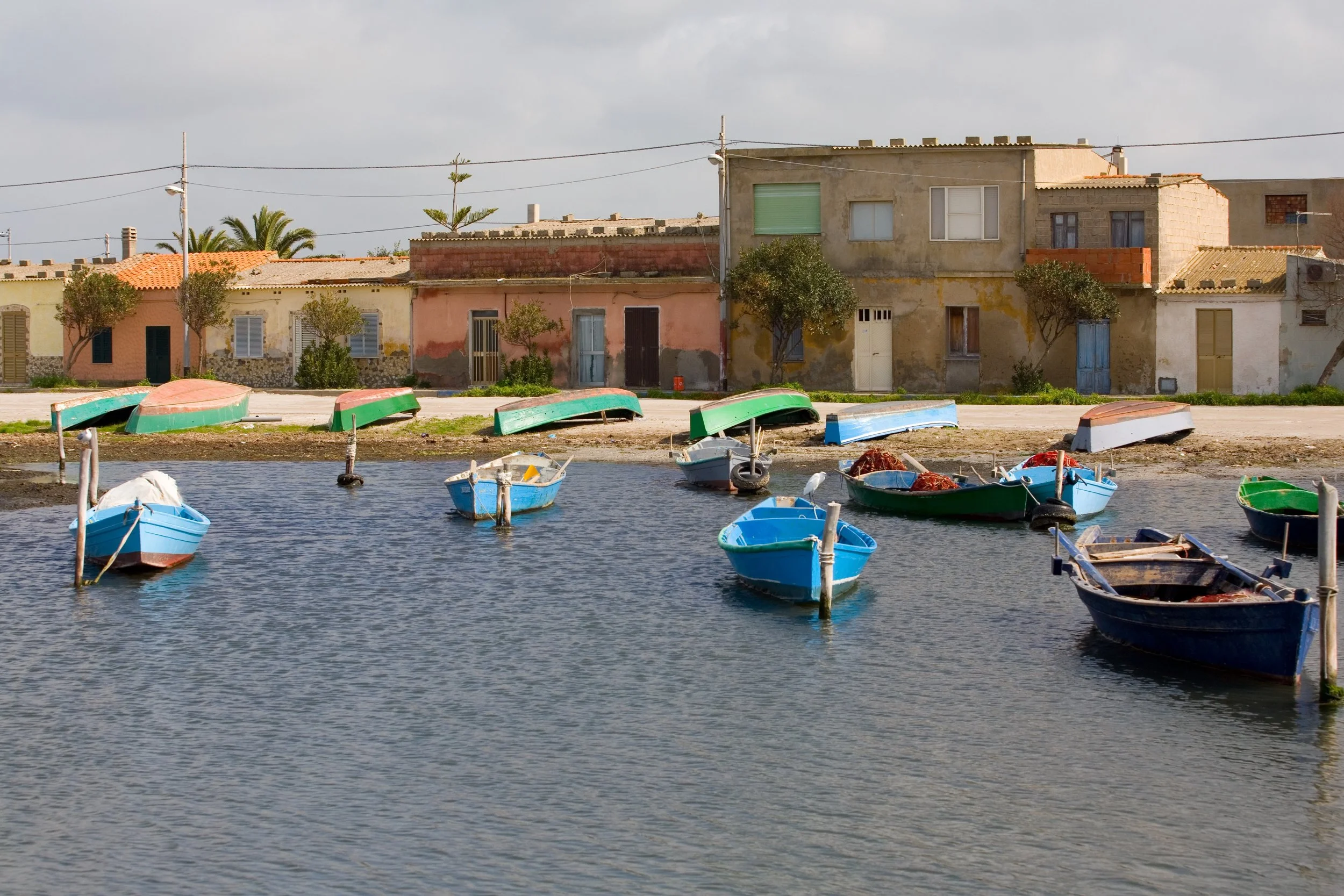 Small fishermen boats anchored in the calm lagoon of Marceddi, Sardinia with a row of colorful, weathered houses and boats on the shore in the background.