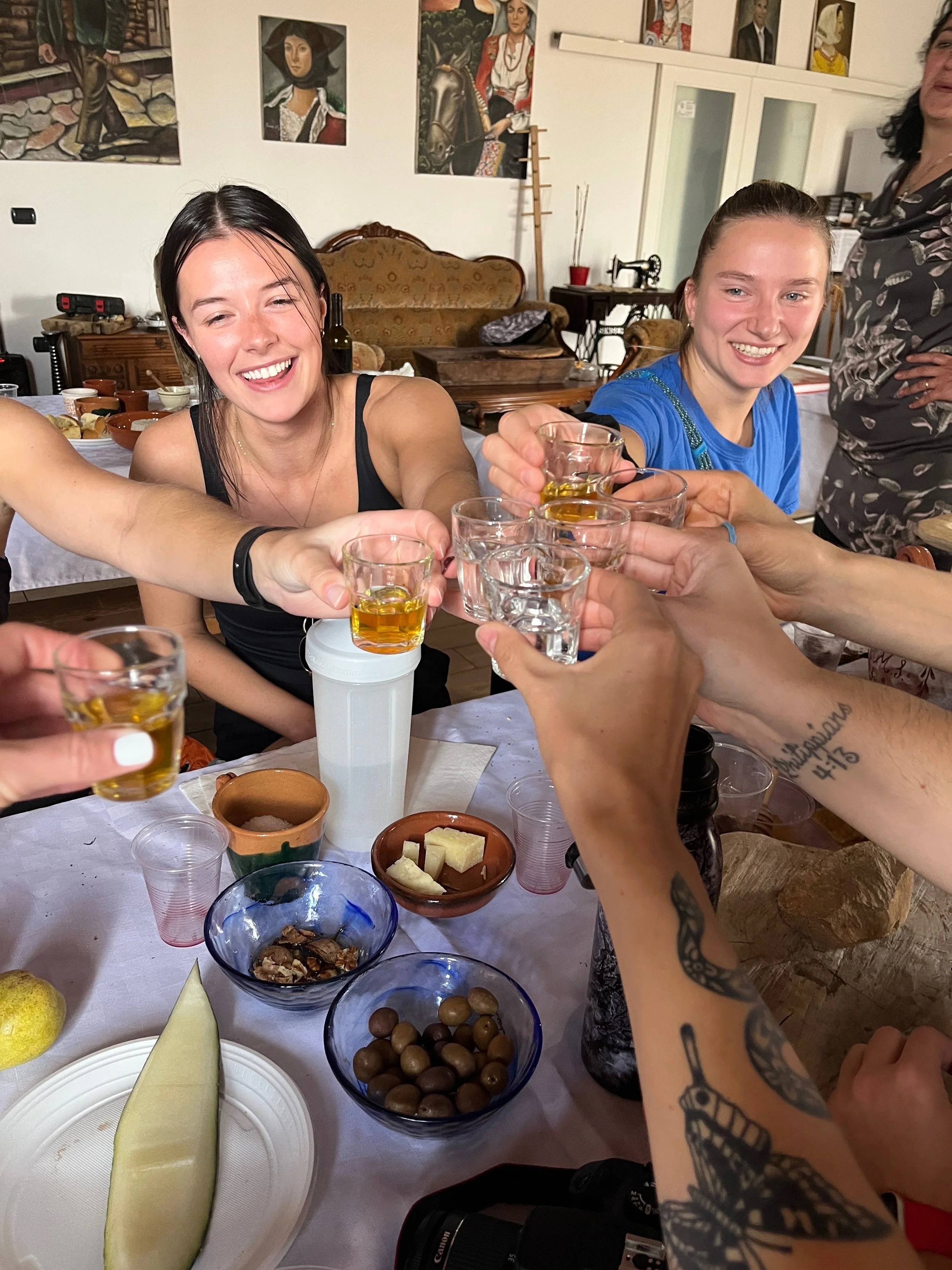 People sitting around a table raising shot glasses filled with a yellowish drink in a toast. The table is set with bowls of snacks, cheese, and fruit. The background includes artwork on the walls and kitchen furniture.