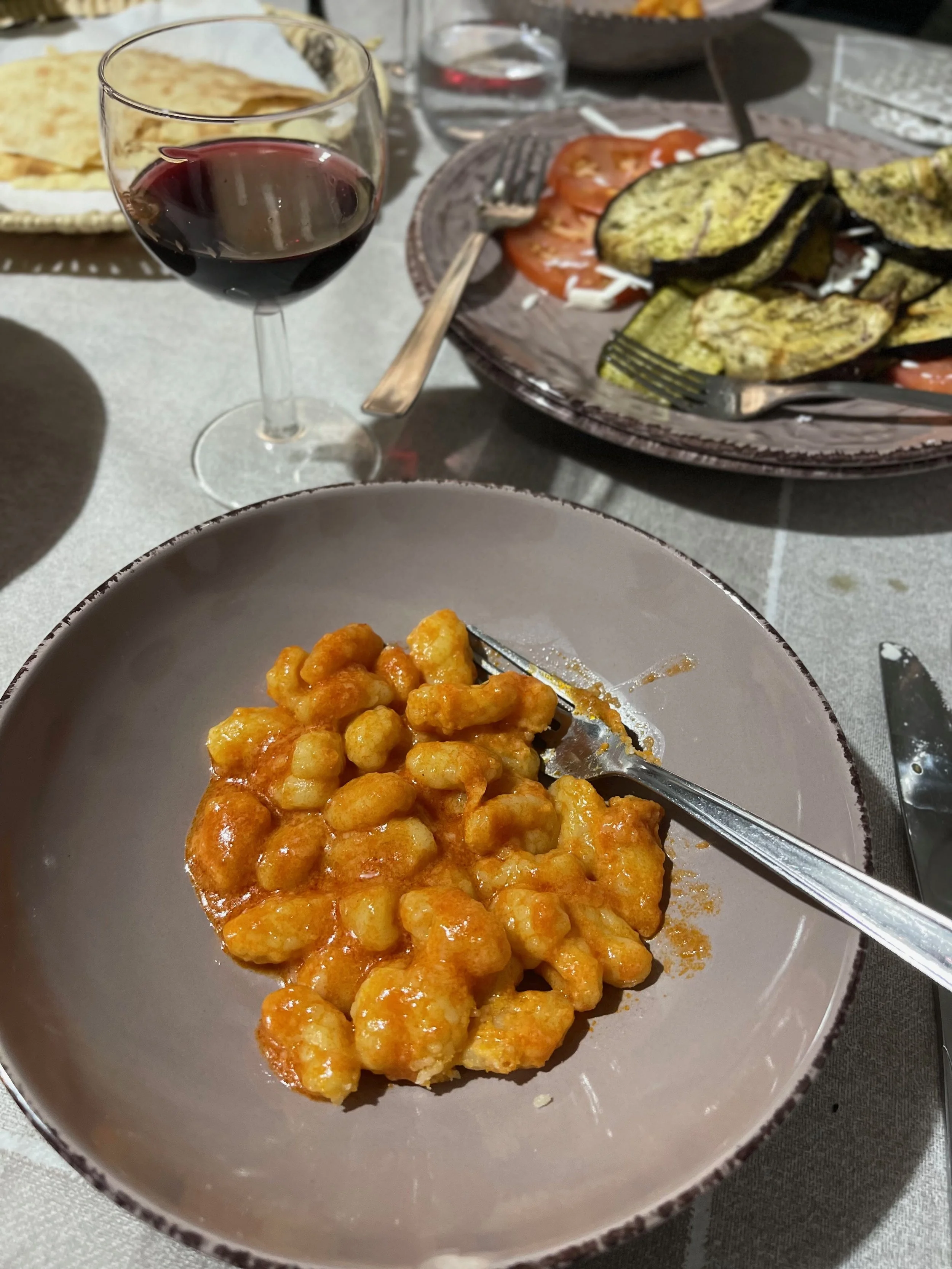 Close-up of a bowl of cheesy fried cauliflower bites on a dining table, with a fork resting inside. In the background, there is a glass of red wine, a plate of grilled vegetables, and a plate of slices of tomato and zucchini.