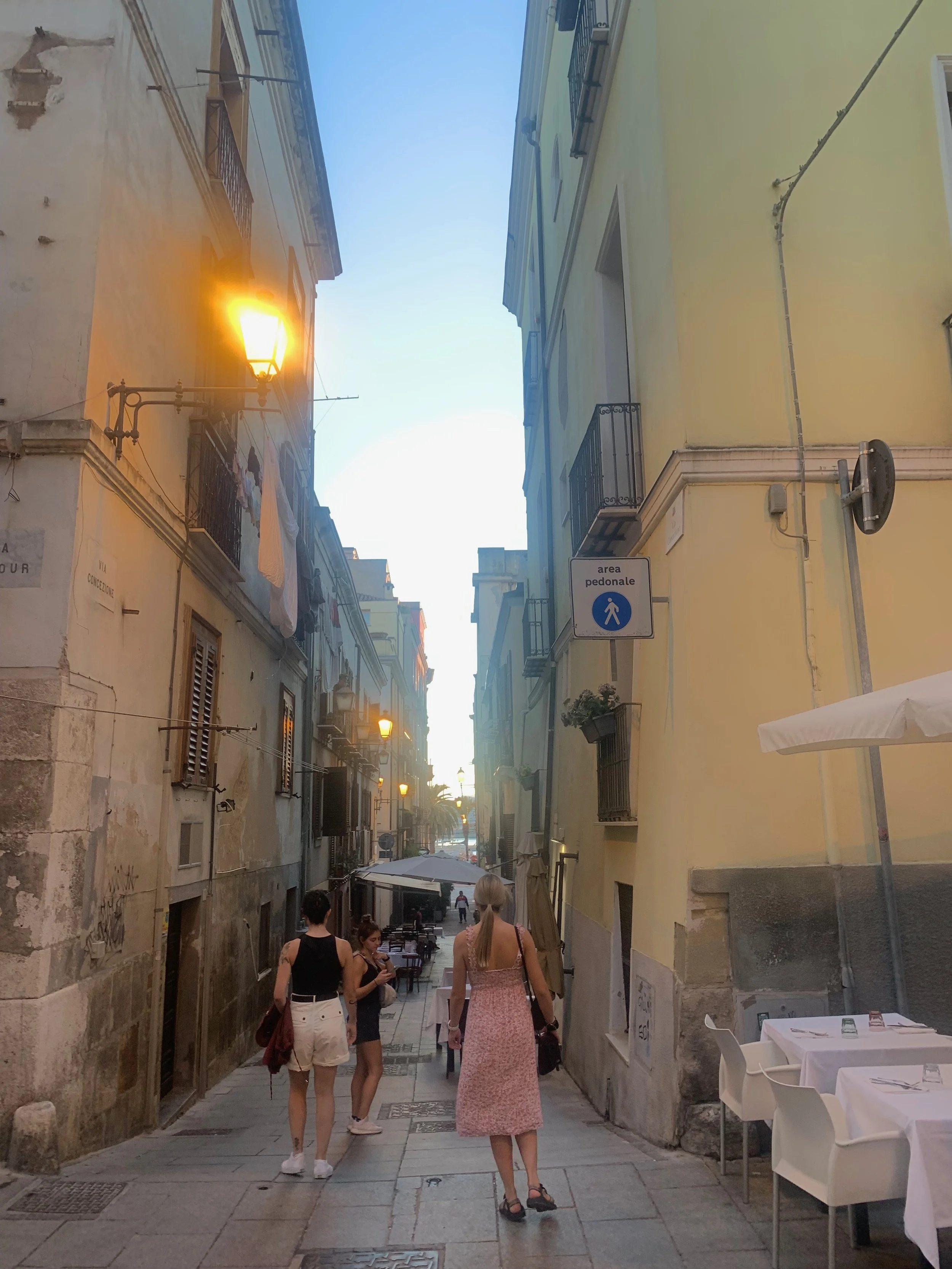 A narrow European street with pastel-colored buildings, outdoor seating, and pedestrians walking during early evening.