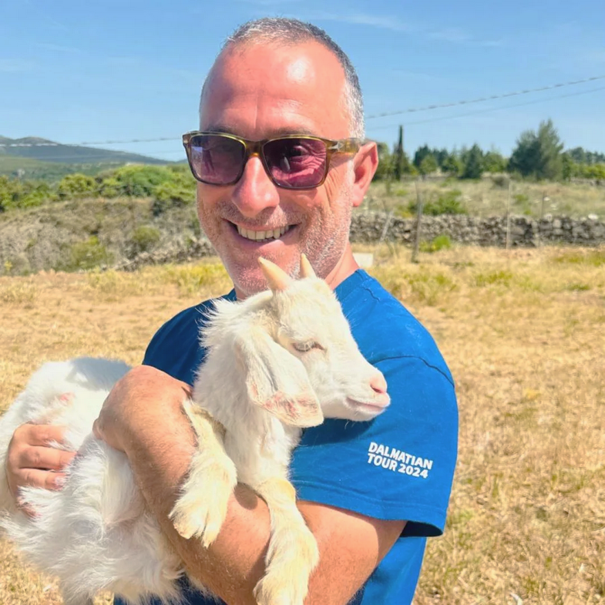 A Sardinia tour guide man in sunglasses smiling and holding a small white goat outdoors with green trees and a stone wall in the background.
