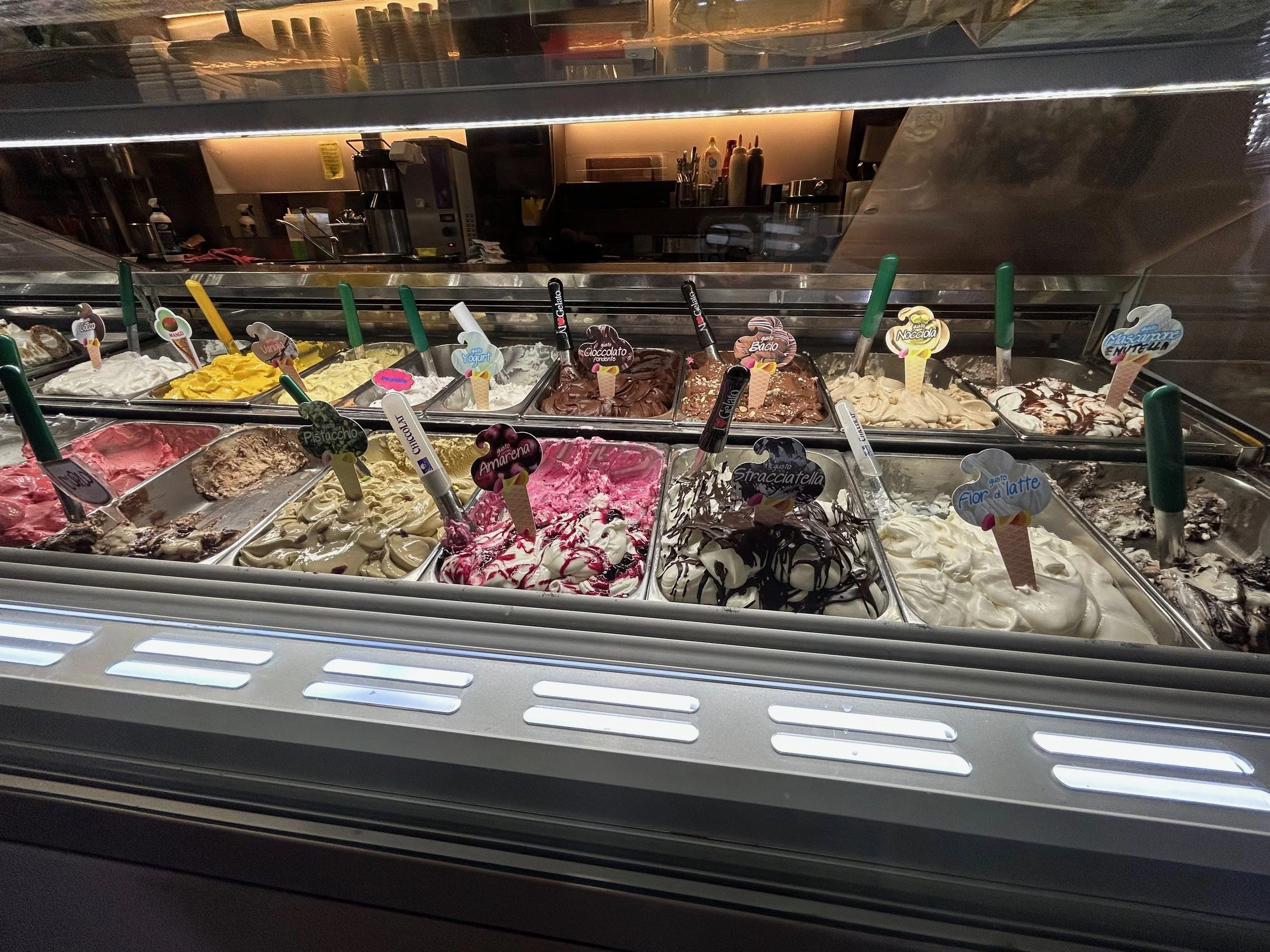Display case of various flavors of gelato with colorful labels and scoops, seen through a glass window in a gelato shop.