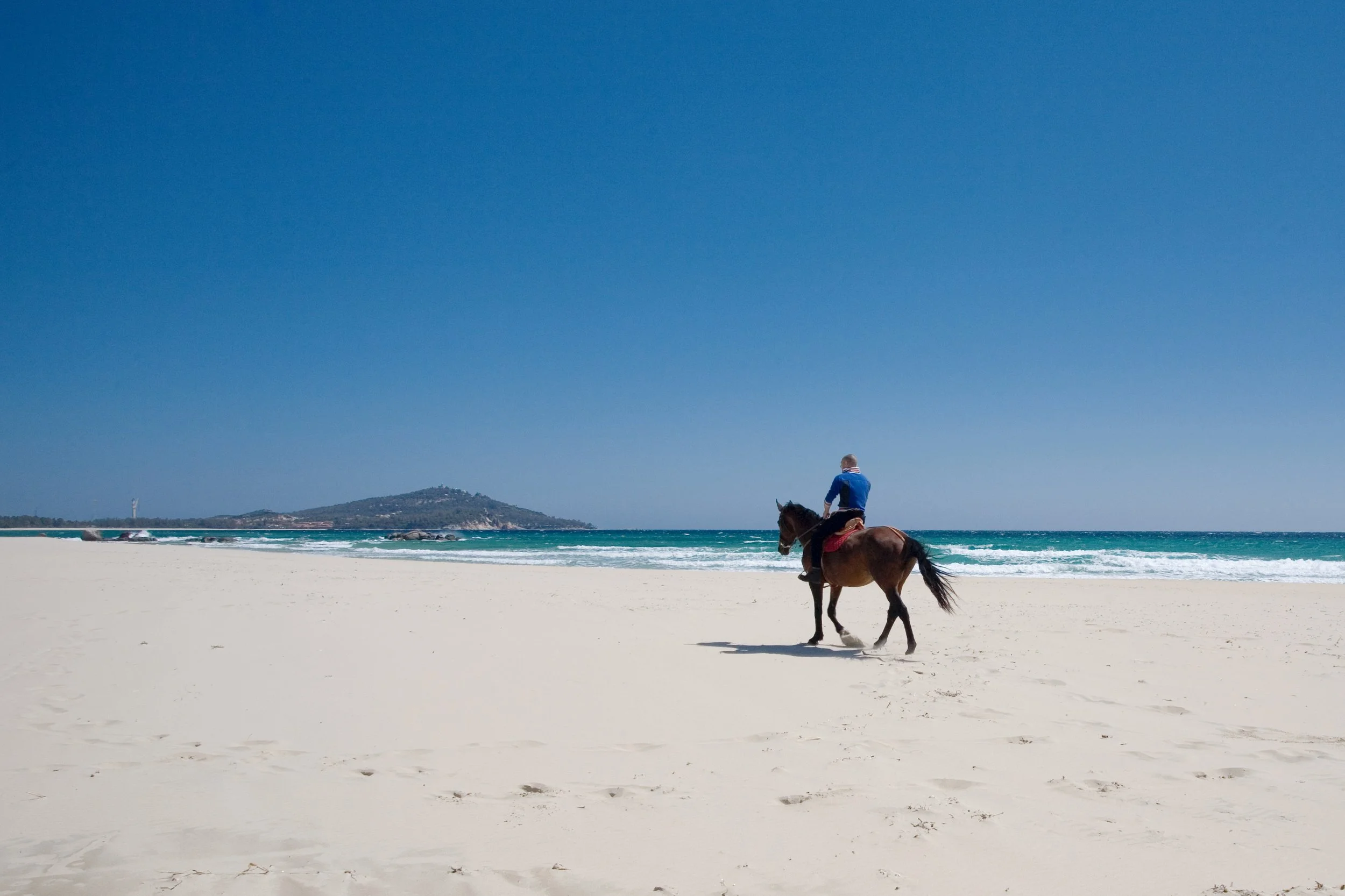 A person riding a horse on a wide sandy beach of Sardinia with the ocean and a hill in the background under a blue sky.