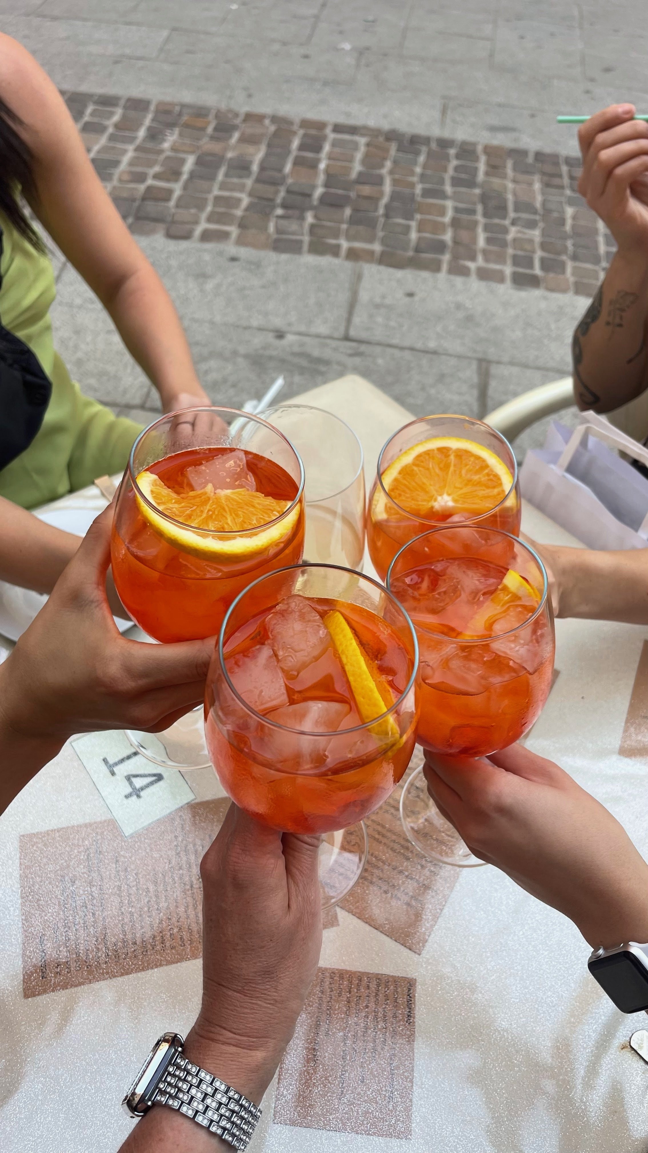 Four glasses of orange-colored drinks with ice and orange slices, being toasted by four people at a table outside.