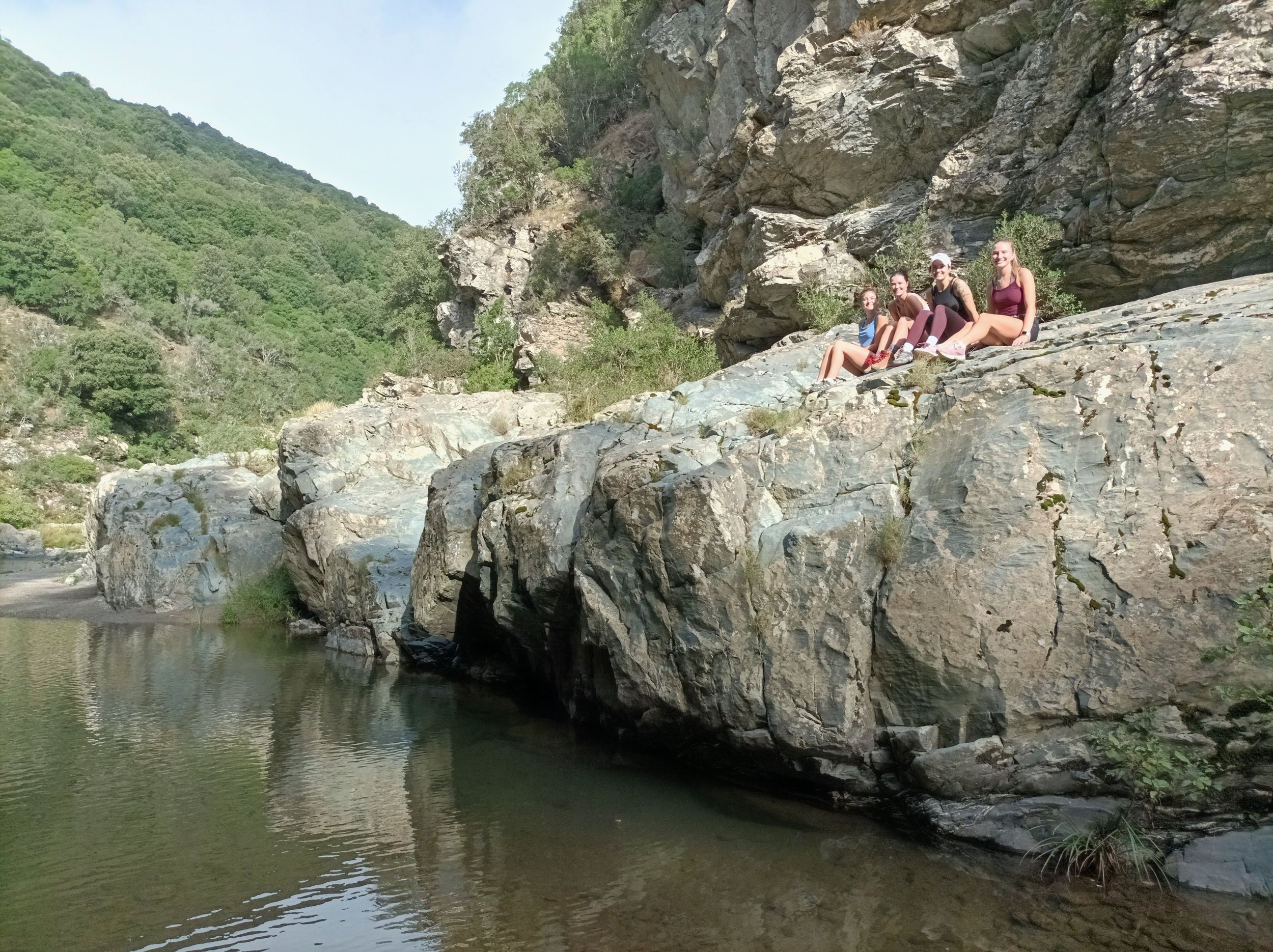 Four young women sitting on large rocks by a river in a canyon with green hills in the background.