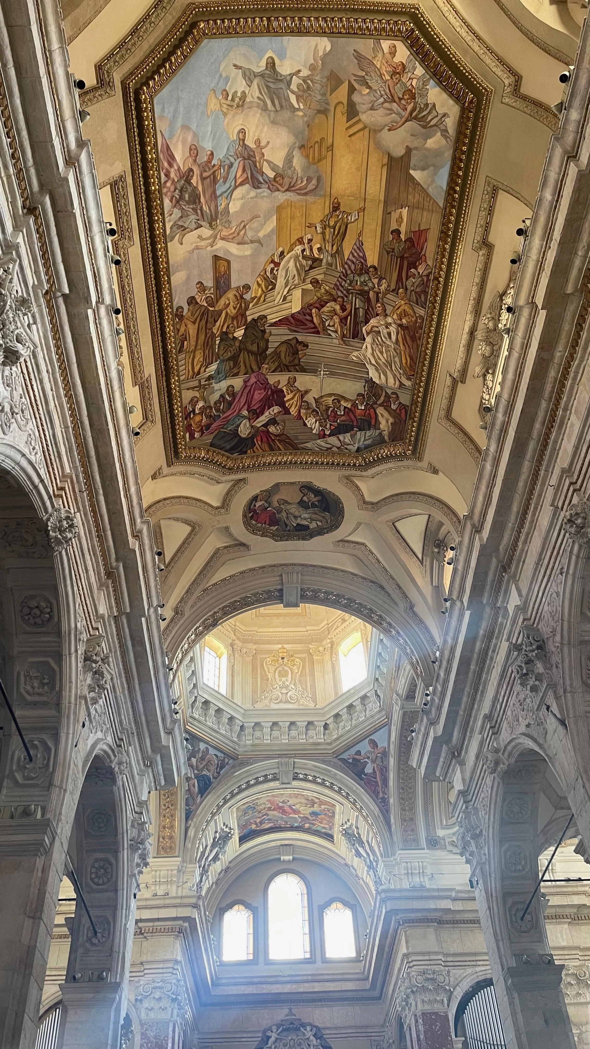 The interior ceiling of a grand church or cathedral featuring ornate stucco work, stained glass windows, and multiple large religious fresco paintings depicting biblical scenes and figures.