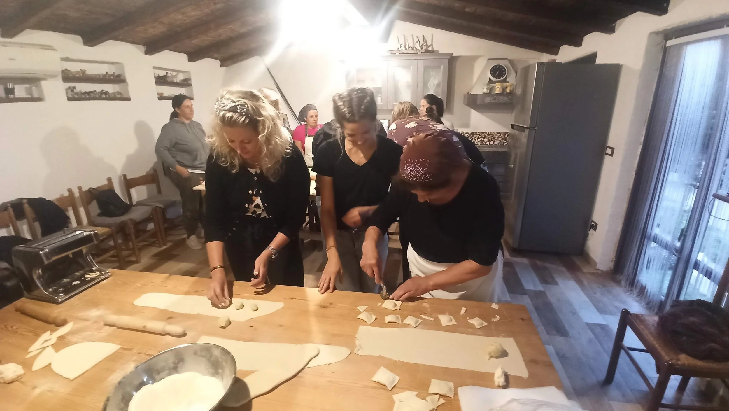 Group of people preparing dough for baking in a kitchen with rustic decor, hooded ceiling, wooden table, and large refrigerator.
