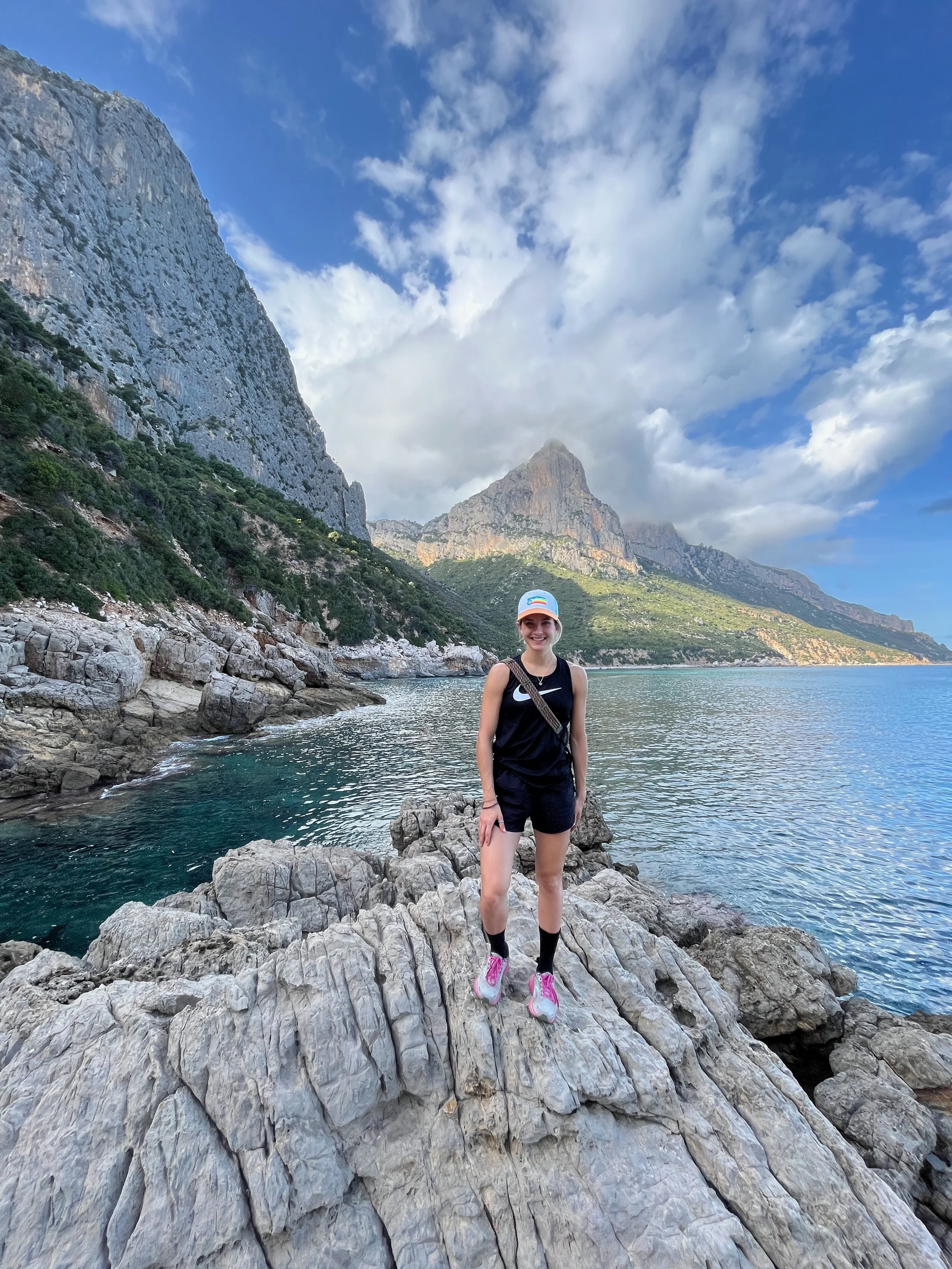 A woman standing on rocks by a body of water with mountains in the background, wearing a black sleeveless top, black shorts, pink and gray sneakers, black socks, a white cap, and a small crossbody bag.