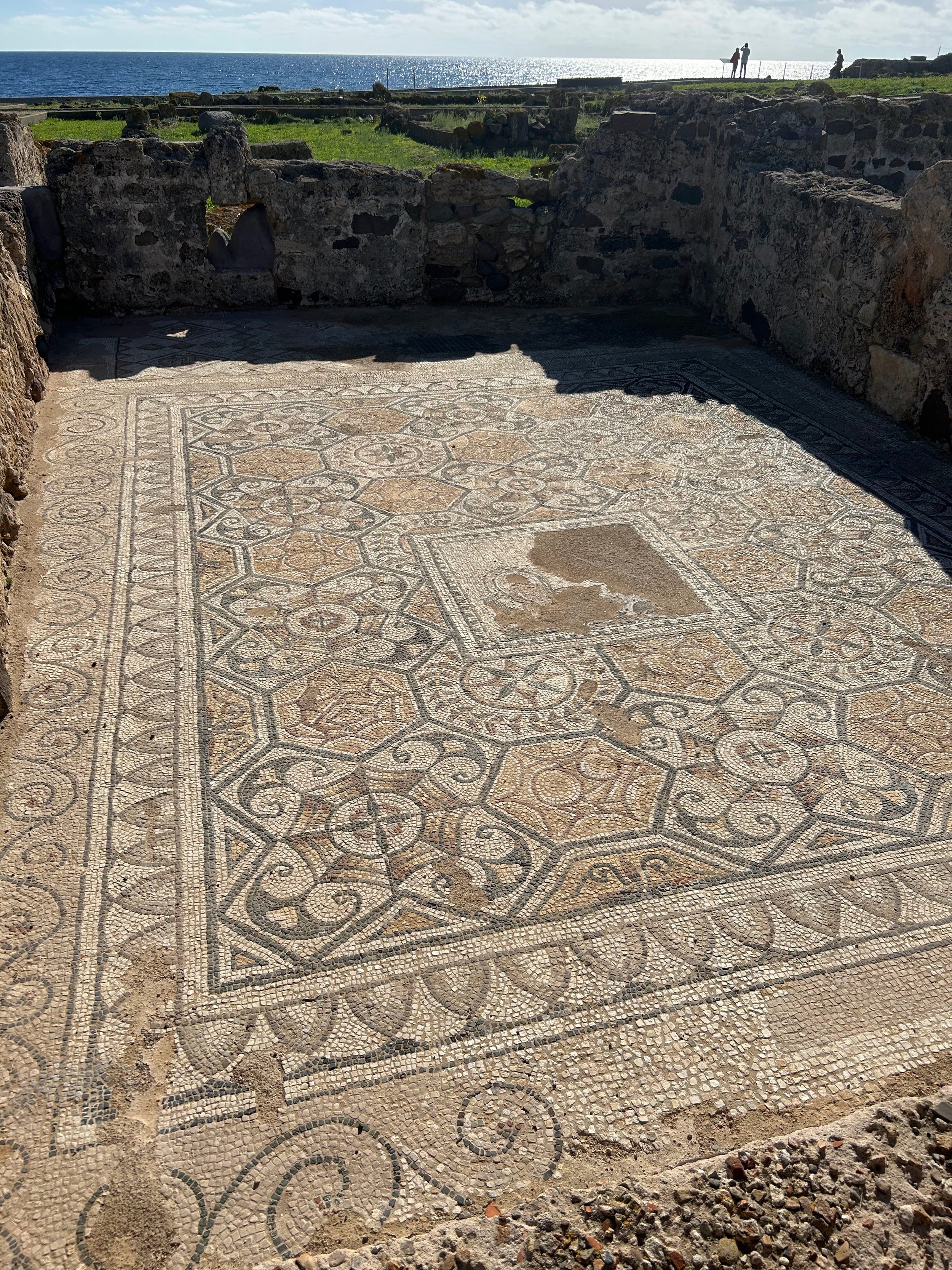 Ancient mosaic floor with geometric and floral patterns, surrounded by stone ruins, overlooking the ocean with visitors in the background.