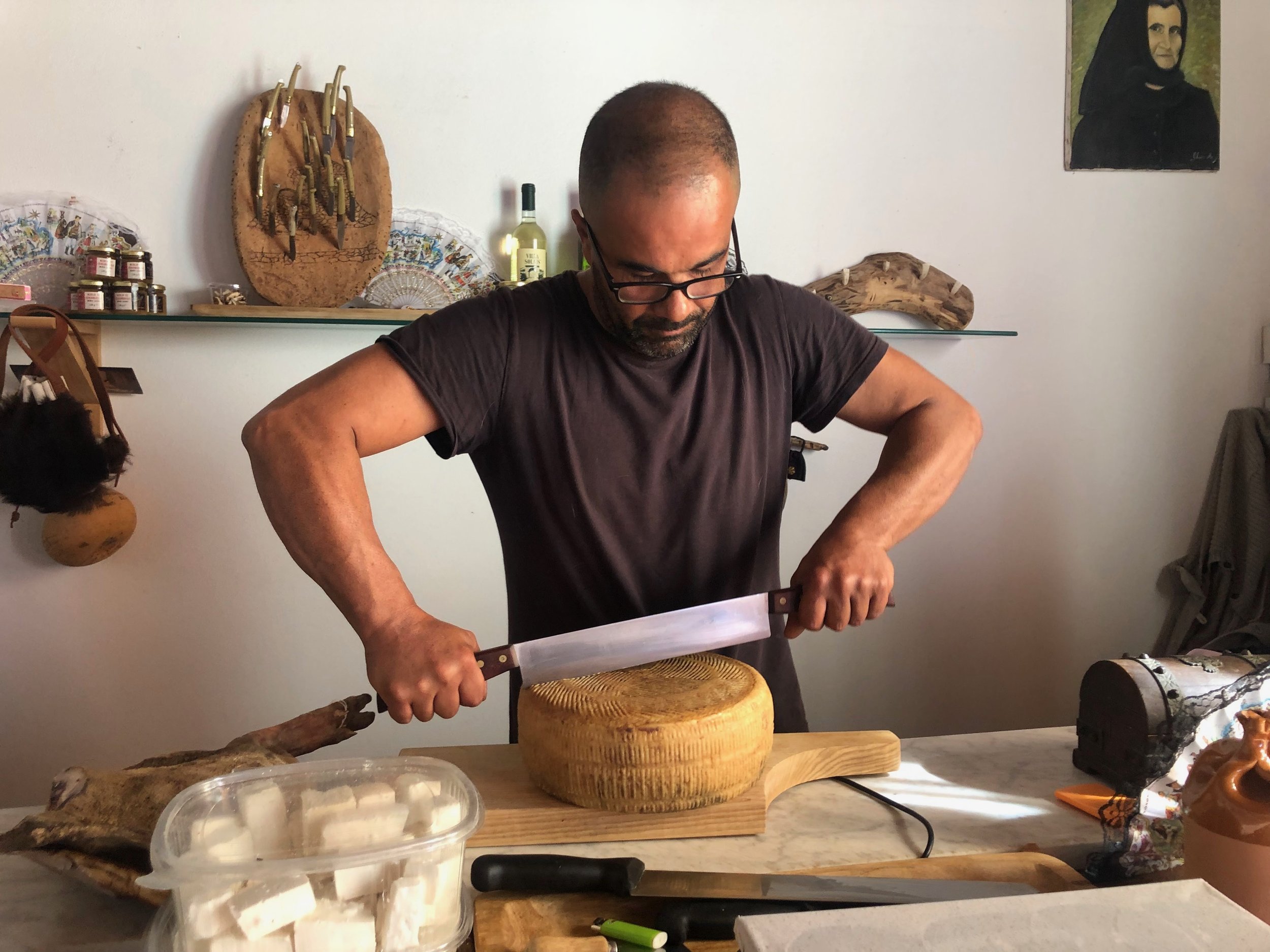 A man wearing glasses and a black shirt is chopping cheese on a wooden cutting board in a kitchen.