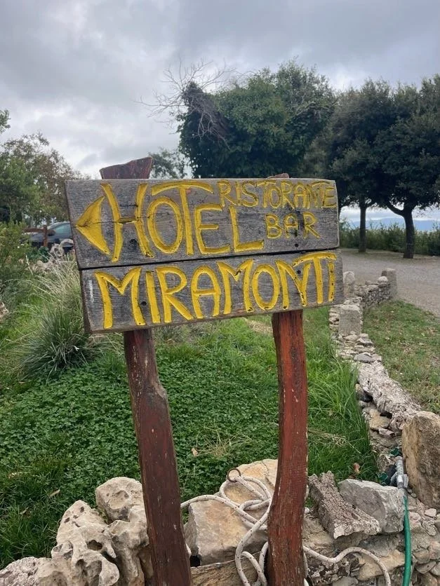 A wooden sign with yellow painted text and an arrow pointing left, advertising a restaurant and bar called Miramont. The sign is outdoors, surrounded by greenery and has a cloudy sky in the background.